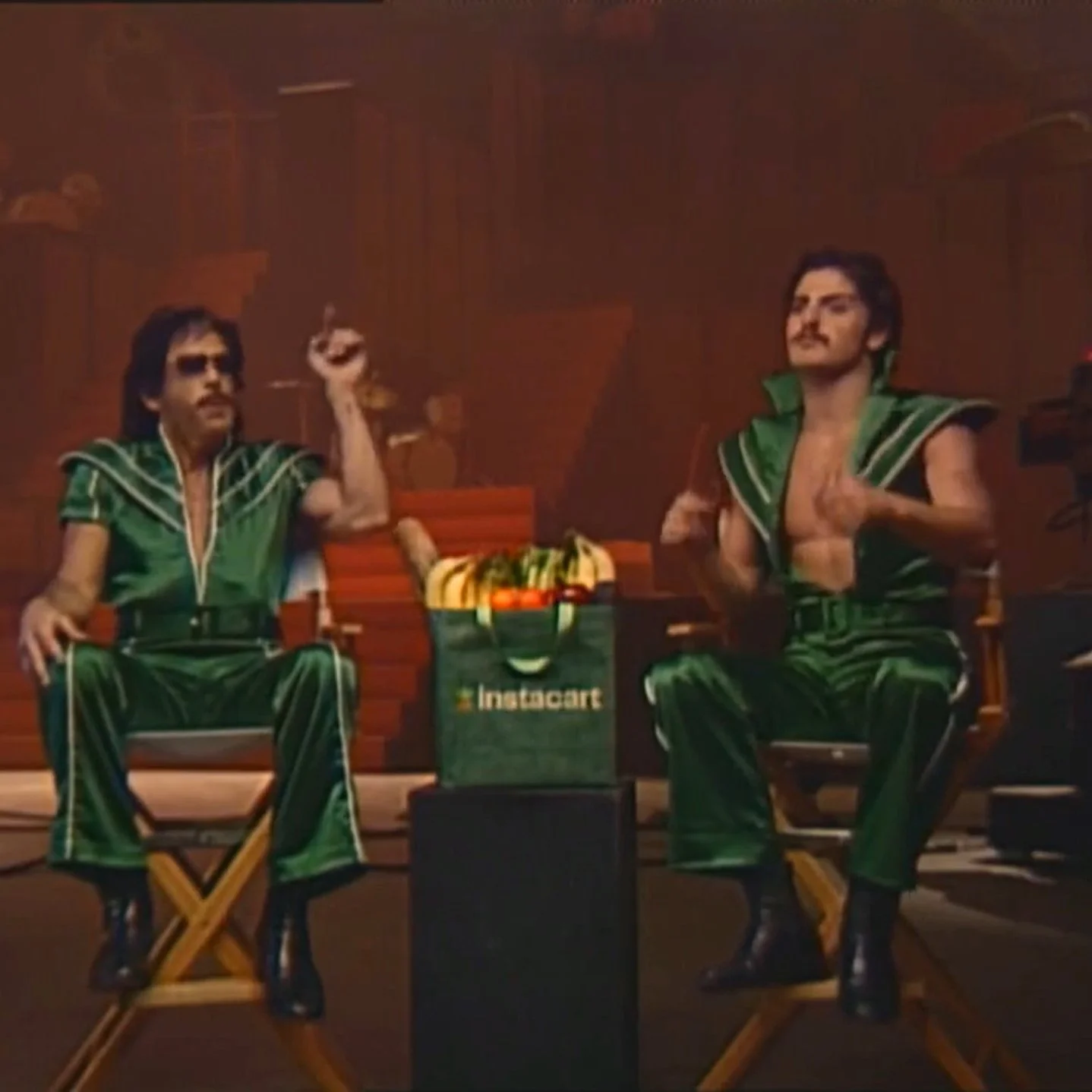 Still image from an instacart commercial showing ben stiller and benson boone dressed in 1970s jumpsuits, sort of like sigfried and roy, sitting in directors chairs, singing