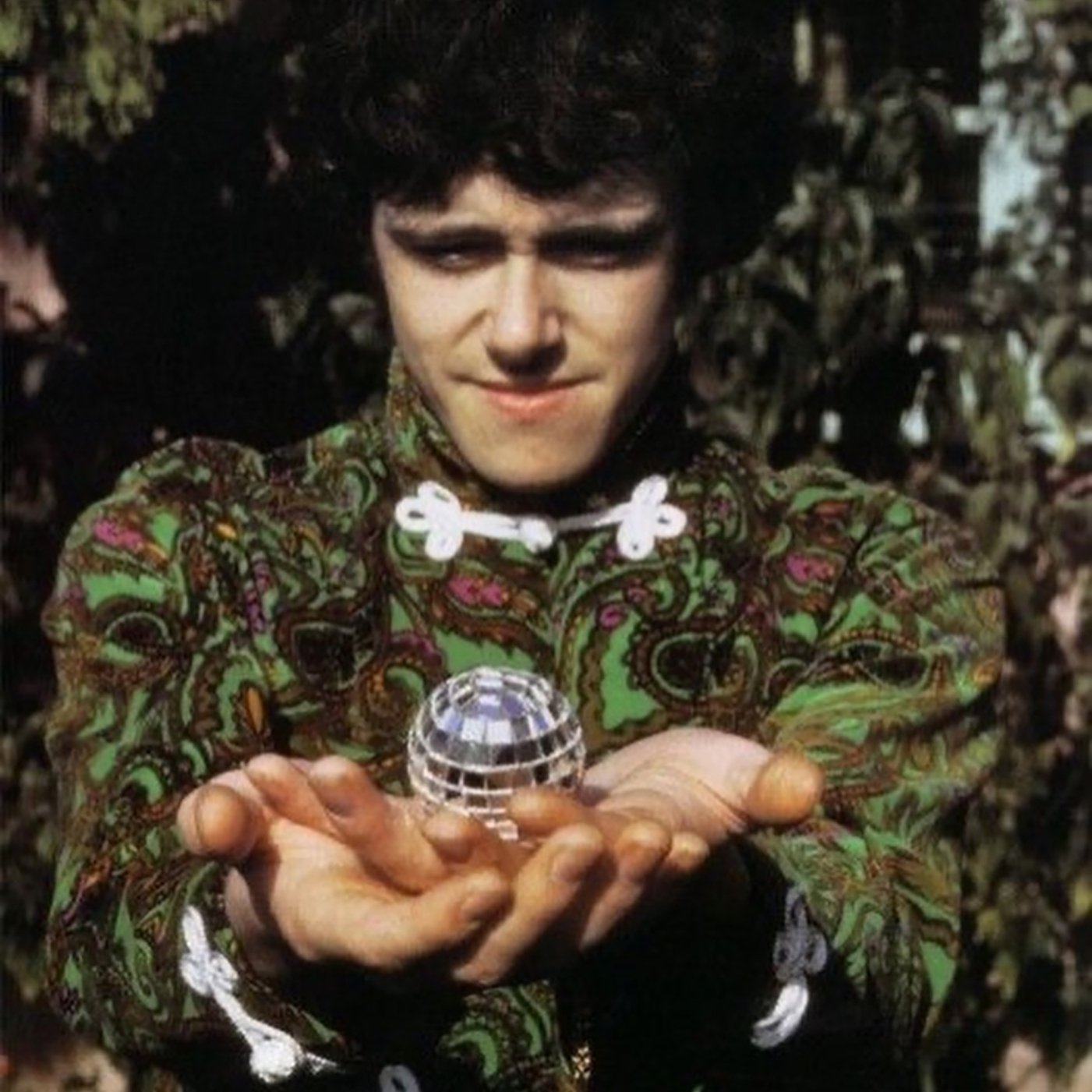 Photo of the singer Donovan in he 1960s dressed in hippie garb and cradling a tiny disco ball in his outstretched hands