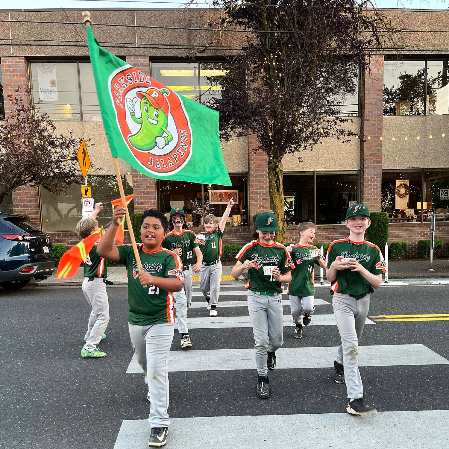 Photo of the TBTL Jr Sluggers little league baseball team crossing the street, carrying a flag with their logo. The kids are in their uniforms.