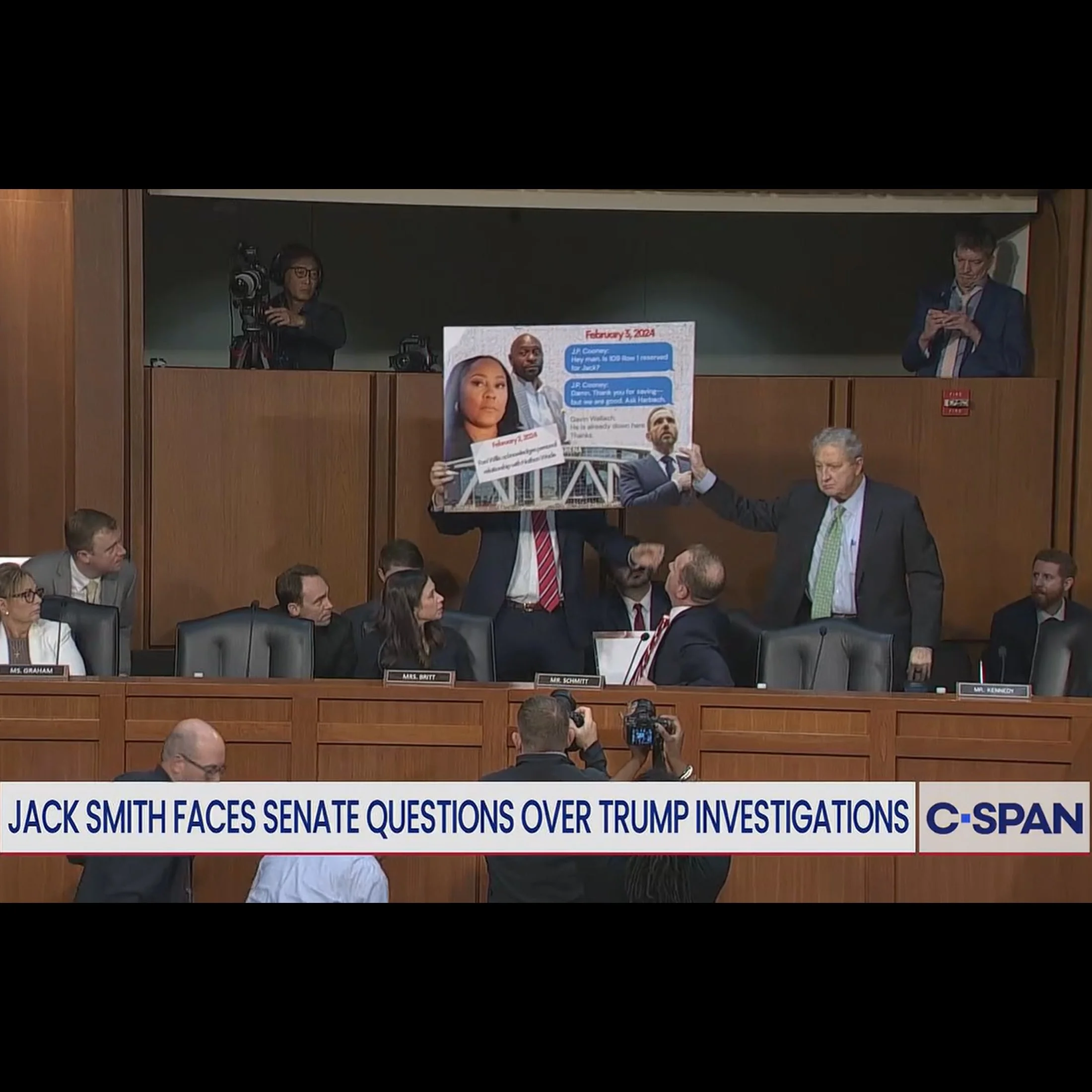 Image from CSPAN showing two men in suits holding up a poster with a janky meme showing text messages and people's faces