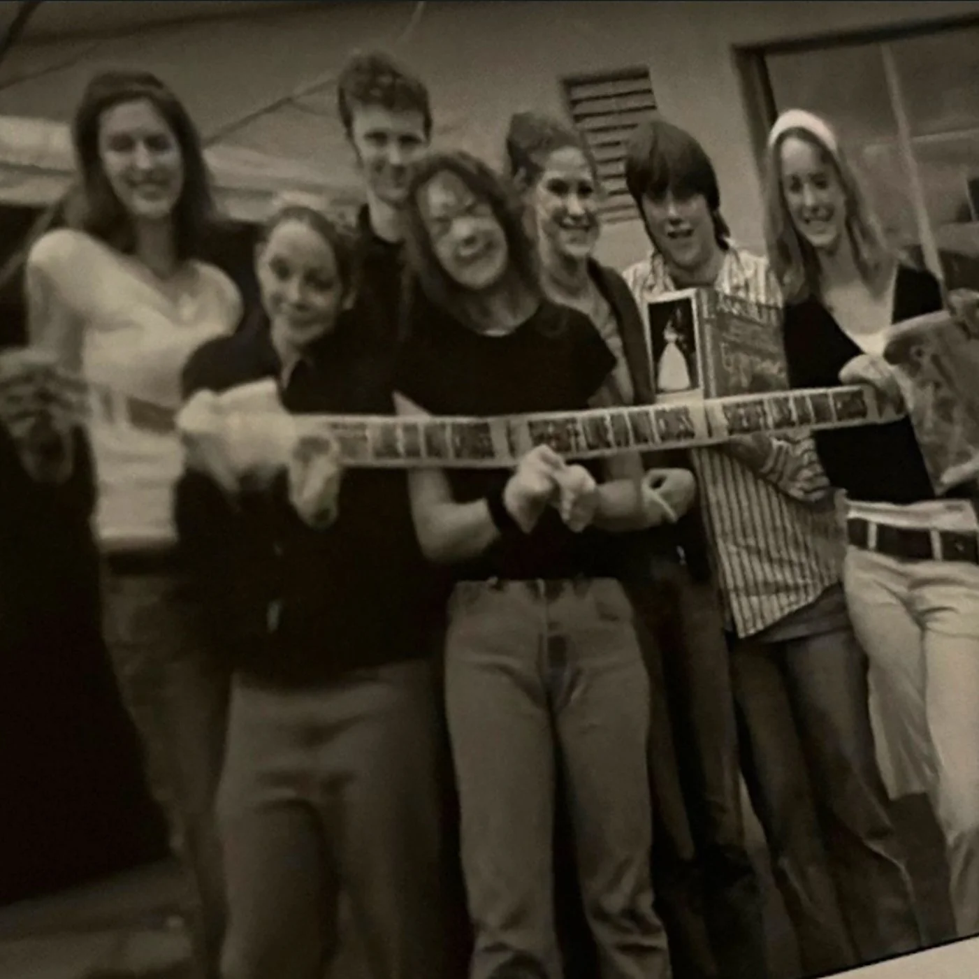 Black and white yearbook photo of a group of kids (including hanna) and an advisor (hanna's mom), posing for a photo. they are rapt in a band of police tape. It's Hanna's Forensic Sciences Club