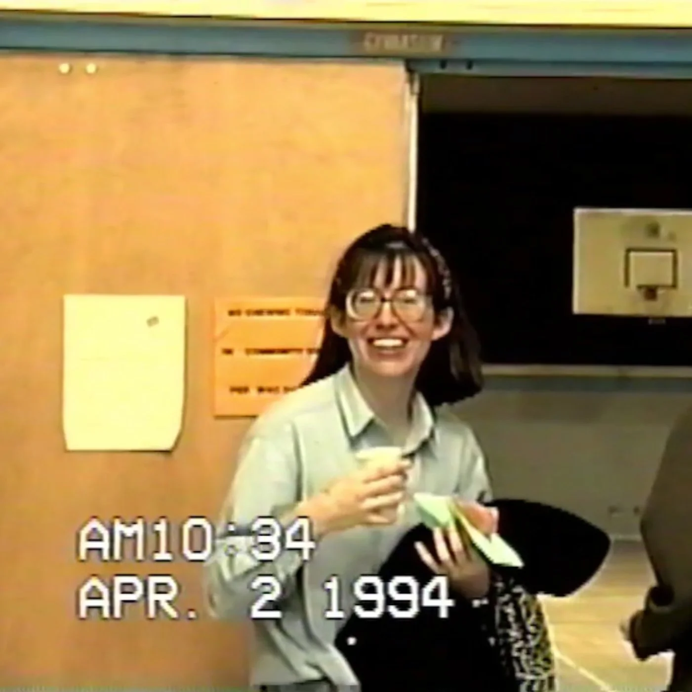 Screengrab from a VHS tape of a woman standing in a doorway in a community center. She's holding a cup of coffee in one hand, a coat and miscellaneous things in her other, as she smiles to the camera. You can see the gymnasium through the door behind