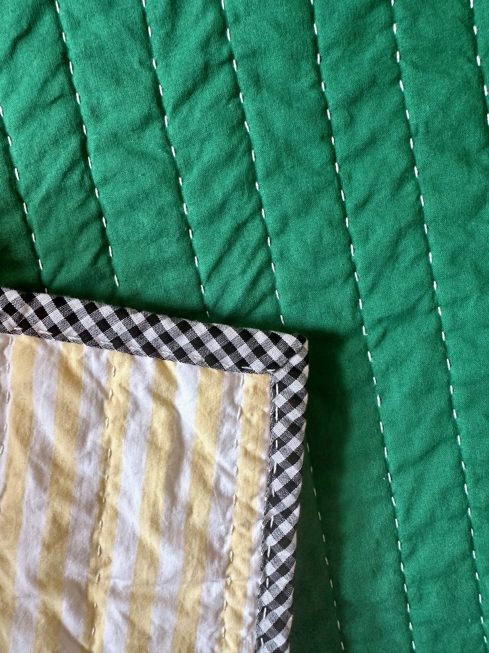 Gingham quilt binding featured against a striped vintage sheet quilt back and a forest green section of patchwork with big stitch hand quilting