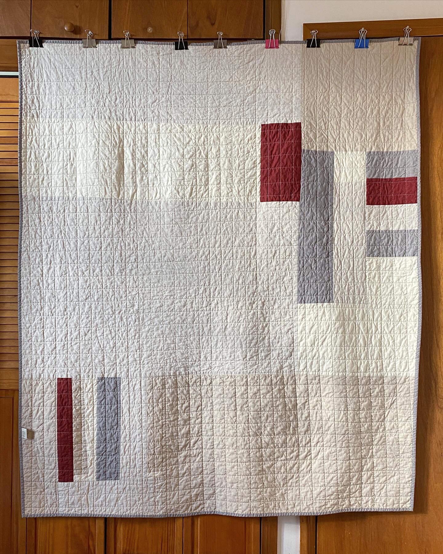 The back of the quilt, hanging straight. Improv-style patchwork composition, made of large rectangular pieces of fabric, in tans, medium gray, and burgundy red.