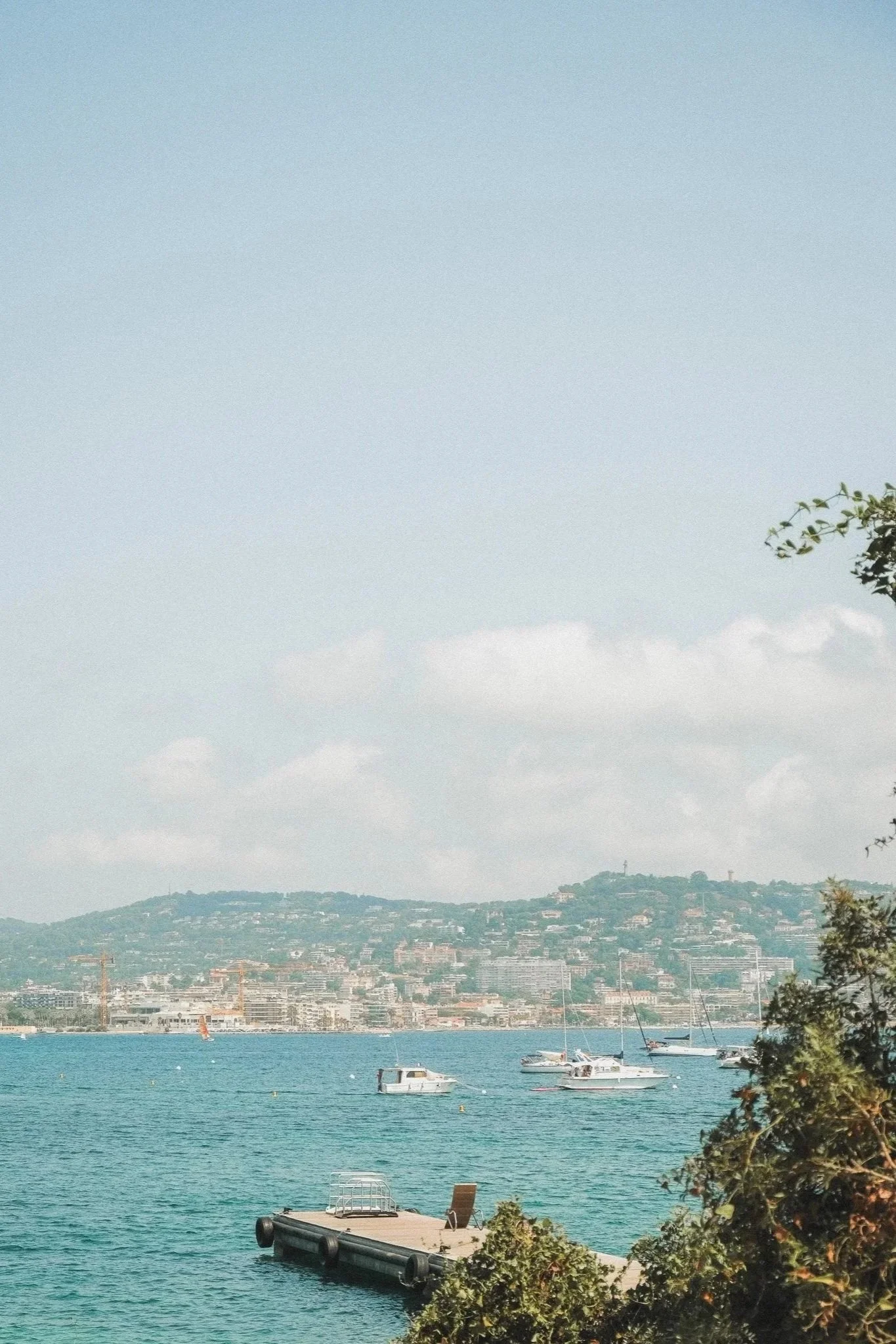 Mountains overlooking a body of water with boats anchored near a pier, and trees in the foreground.
