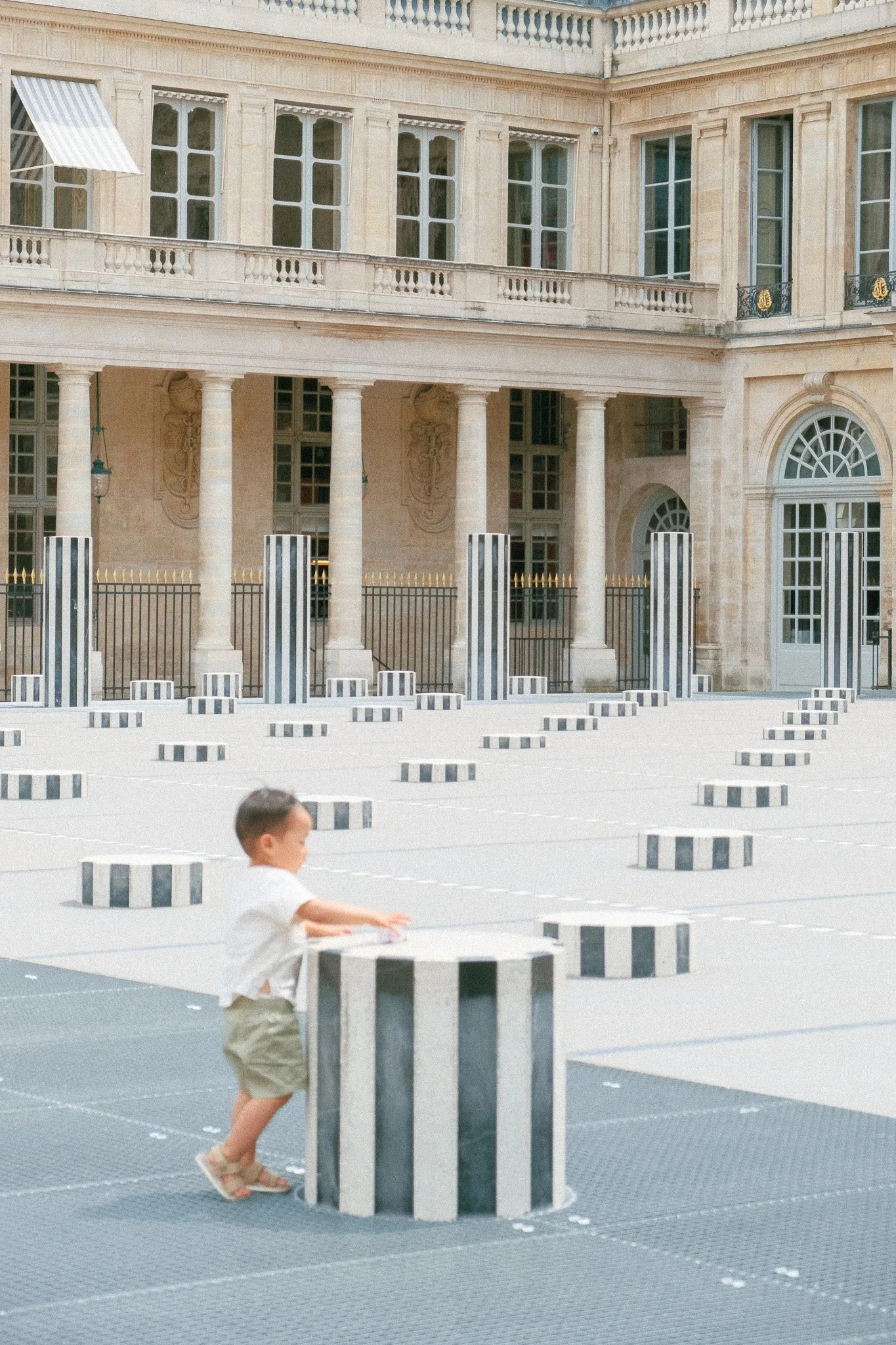 A young boy in a white shirt and shorts interacts with striped black and white cylindrical sculptures in an outdoor urban plaza with classical architecture, large windows, and columns.