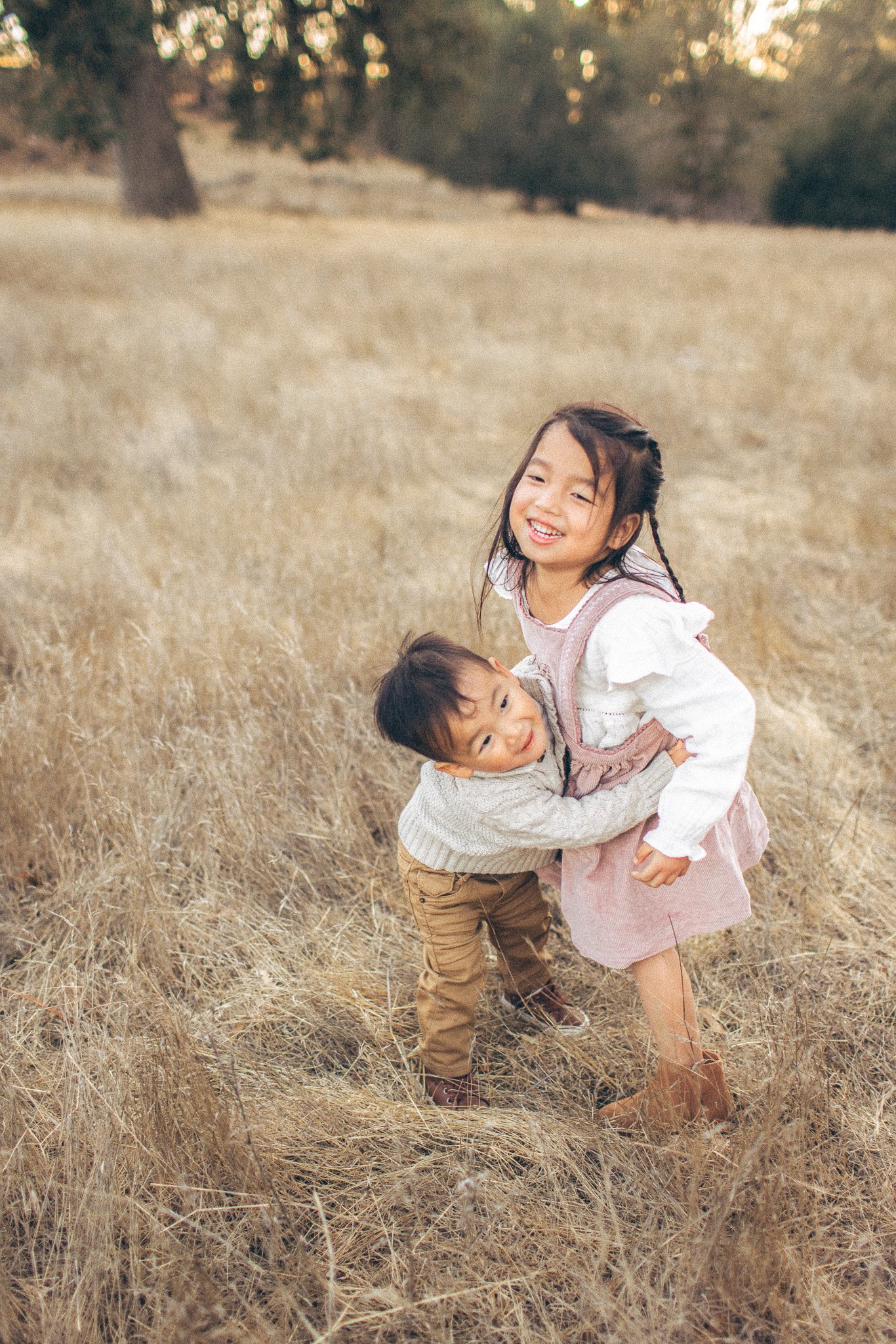 A girl and a boy playing and hugging in a dry, grassy field with trees in the background.