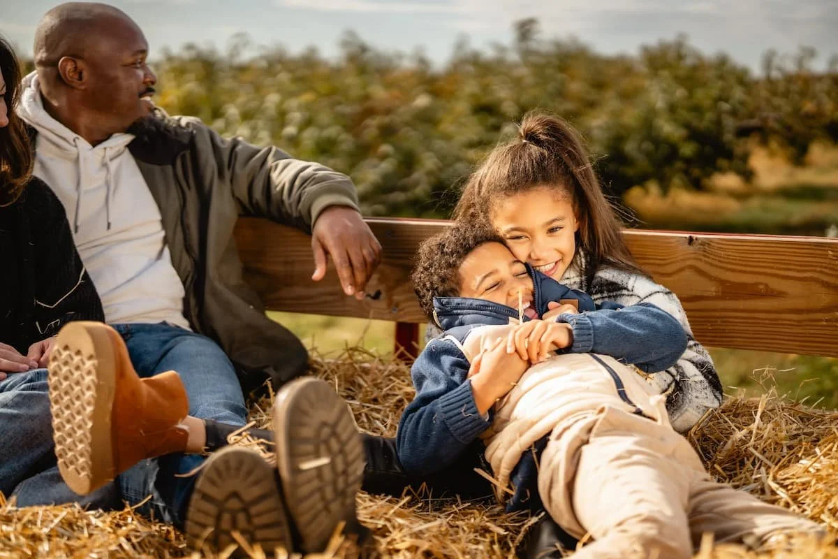 A family sitting on a bed of hay outdoors, with children laughing and hugging, under a wooden fence, with trees in the background and a partly cloudy sky.