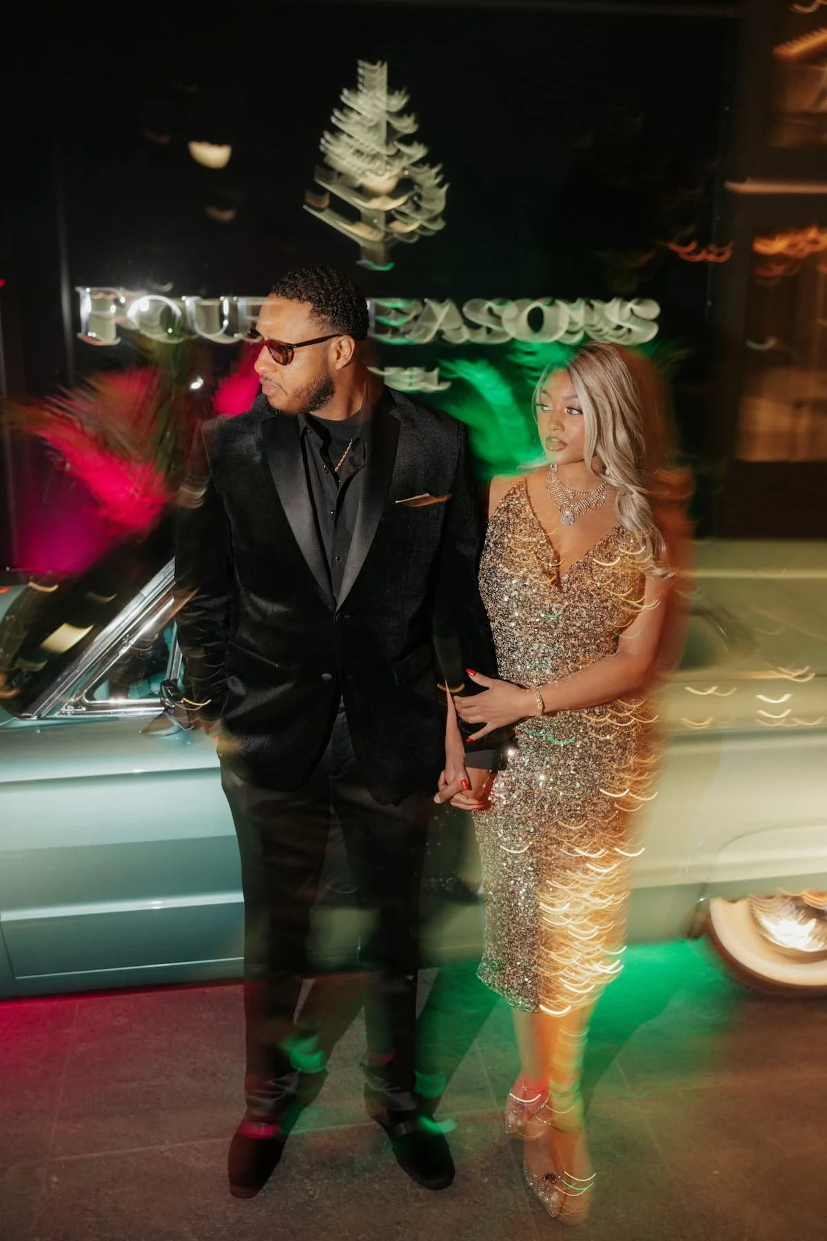 A man in a black velvet suit and sunglasses standing next to a woman in a sparkling gold dress outside a building at night.