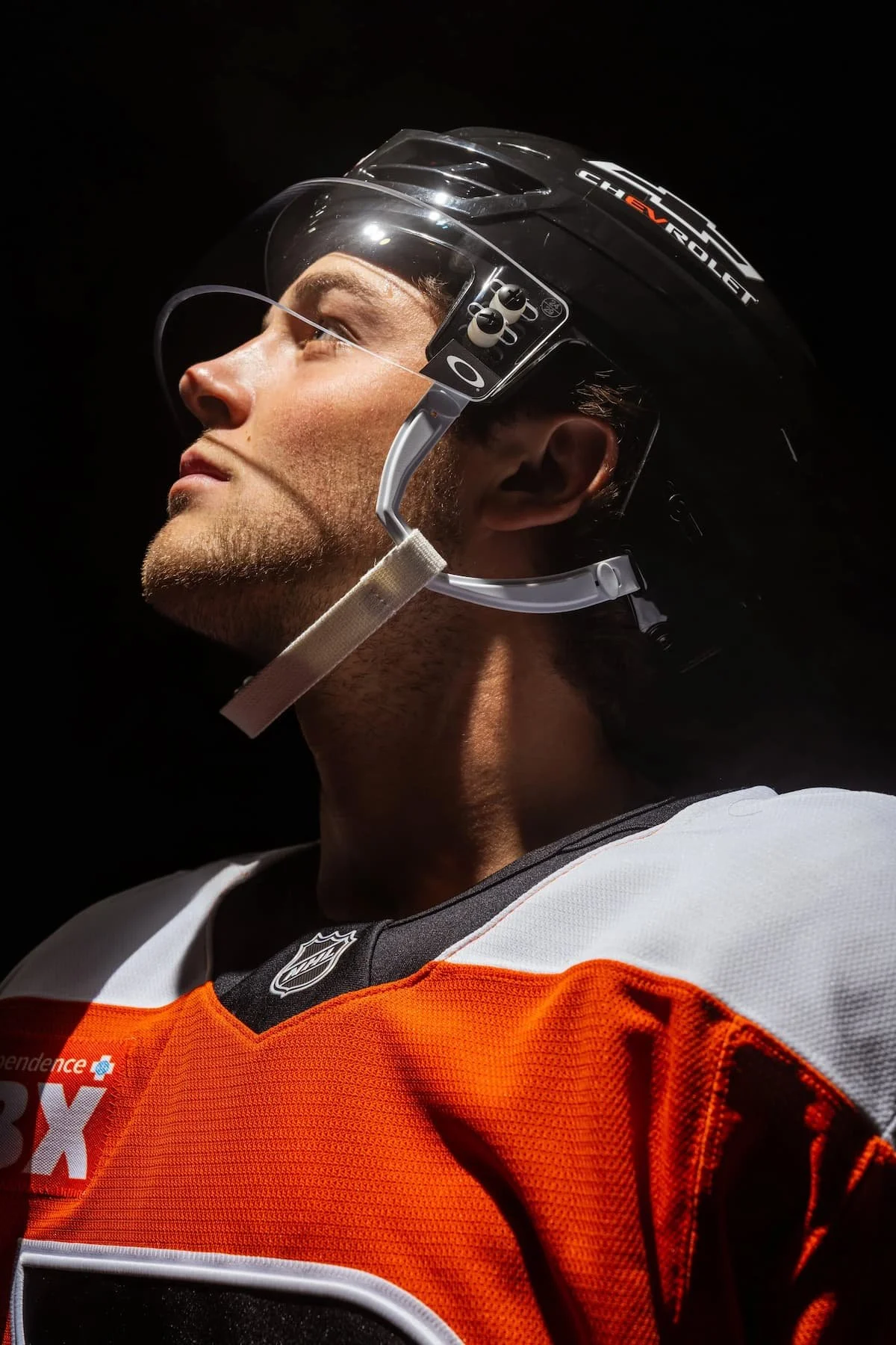 A Philadelphia Flyer hockey player in a black helmet with a visor and orange and black jersey, looking upward against a dark background.