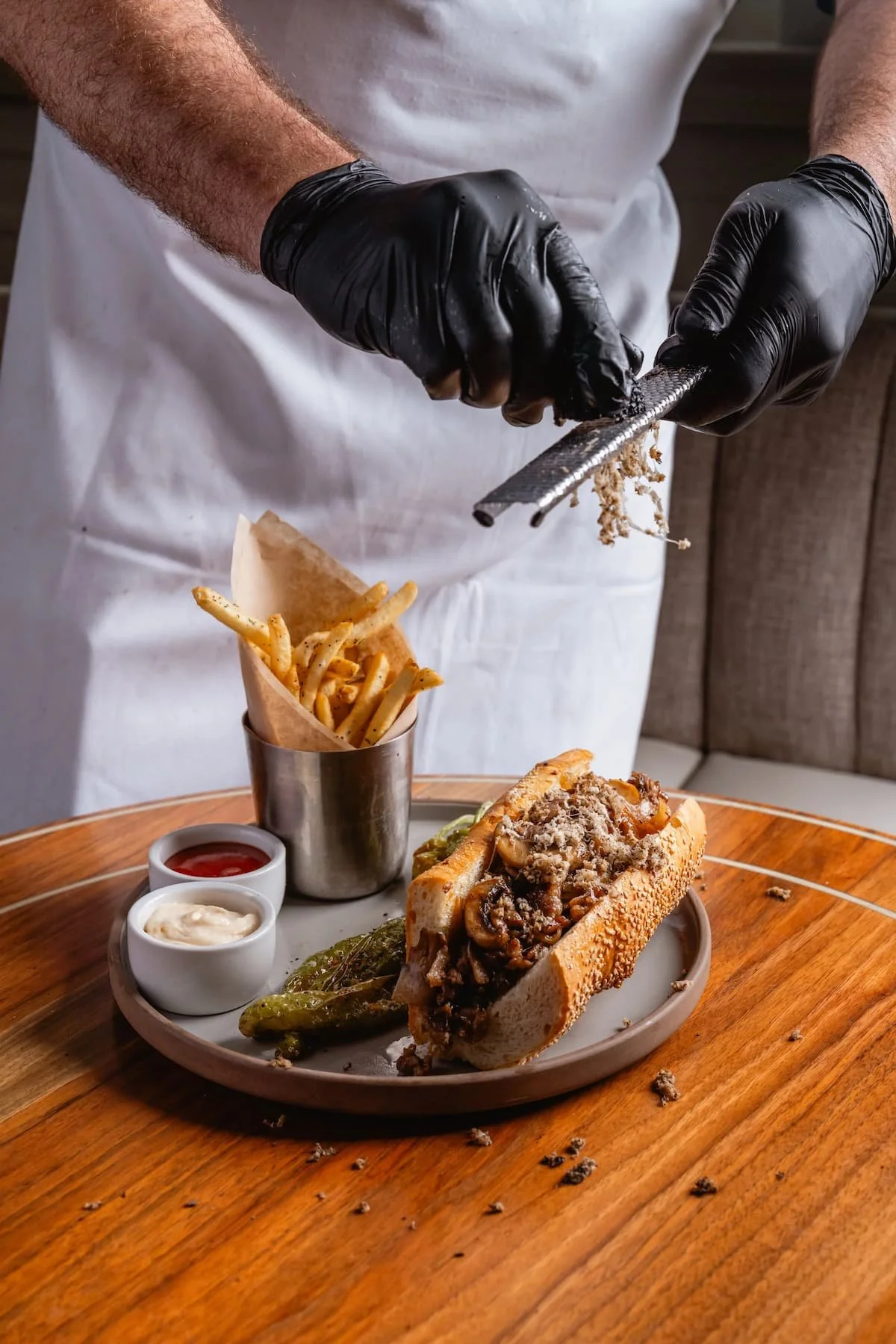 Chef wearing black gloves grating cheese over a sausage sandwich with fries and condiments on a round wooden table