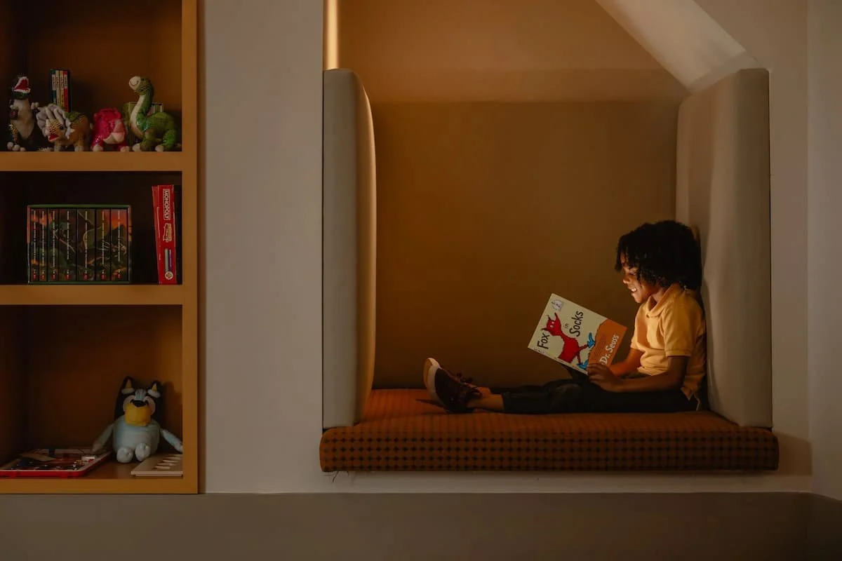 A young boy with curly hair sitting in a cozy nook reading a Dr. Seuss book titled "Fox in Socks" with a smile, surrounded by toys and books on a shelf nearby.
