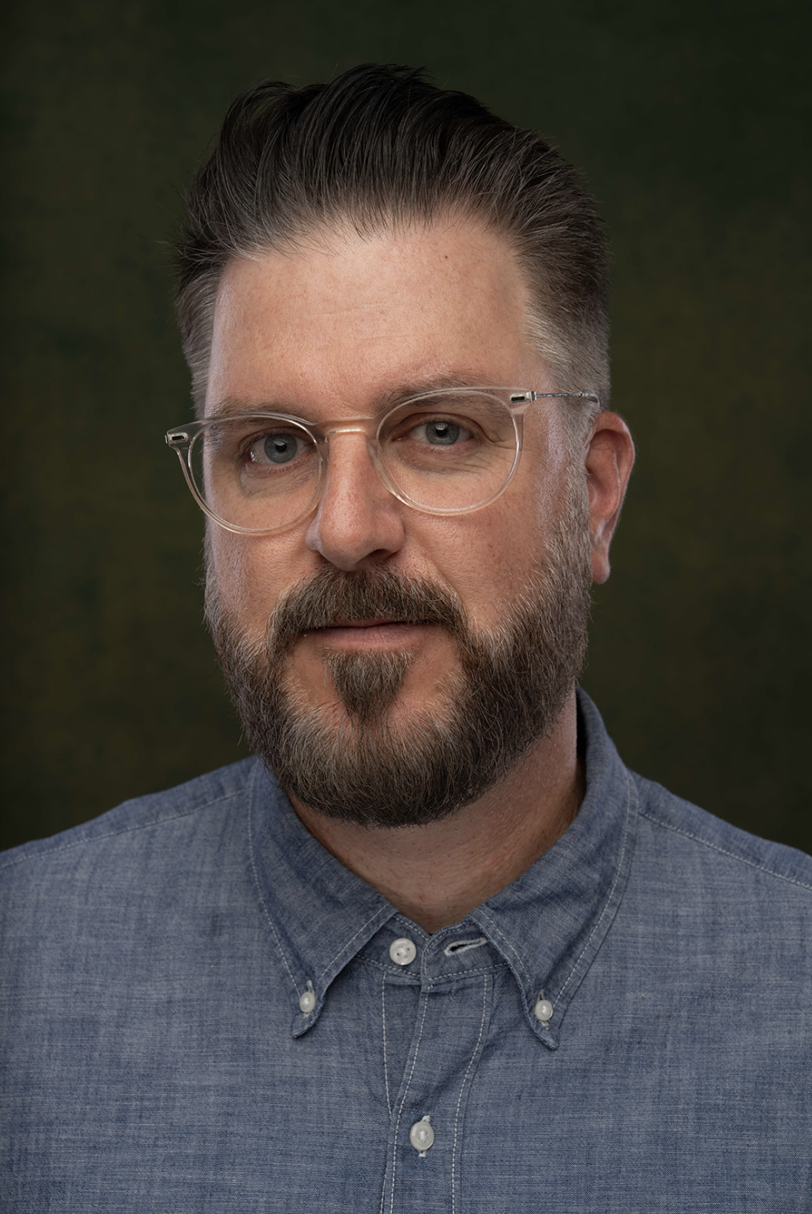 Close-up portrait of a man with glasses and a beard wearing a denim button-up shirt, against a dark green background.