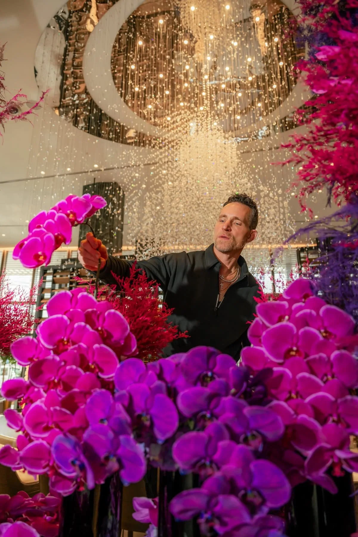A man arranging pink orchids and red flowers in a floral display with a backdrop of hanging string lights and a reflective ceiling.