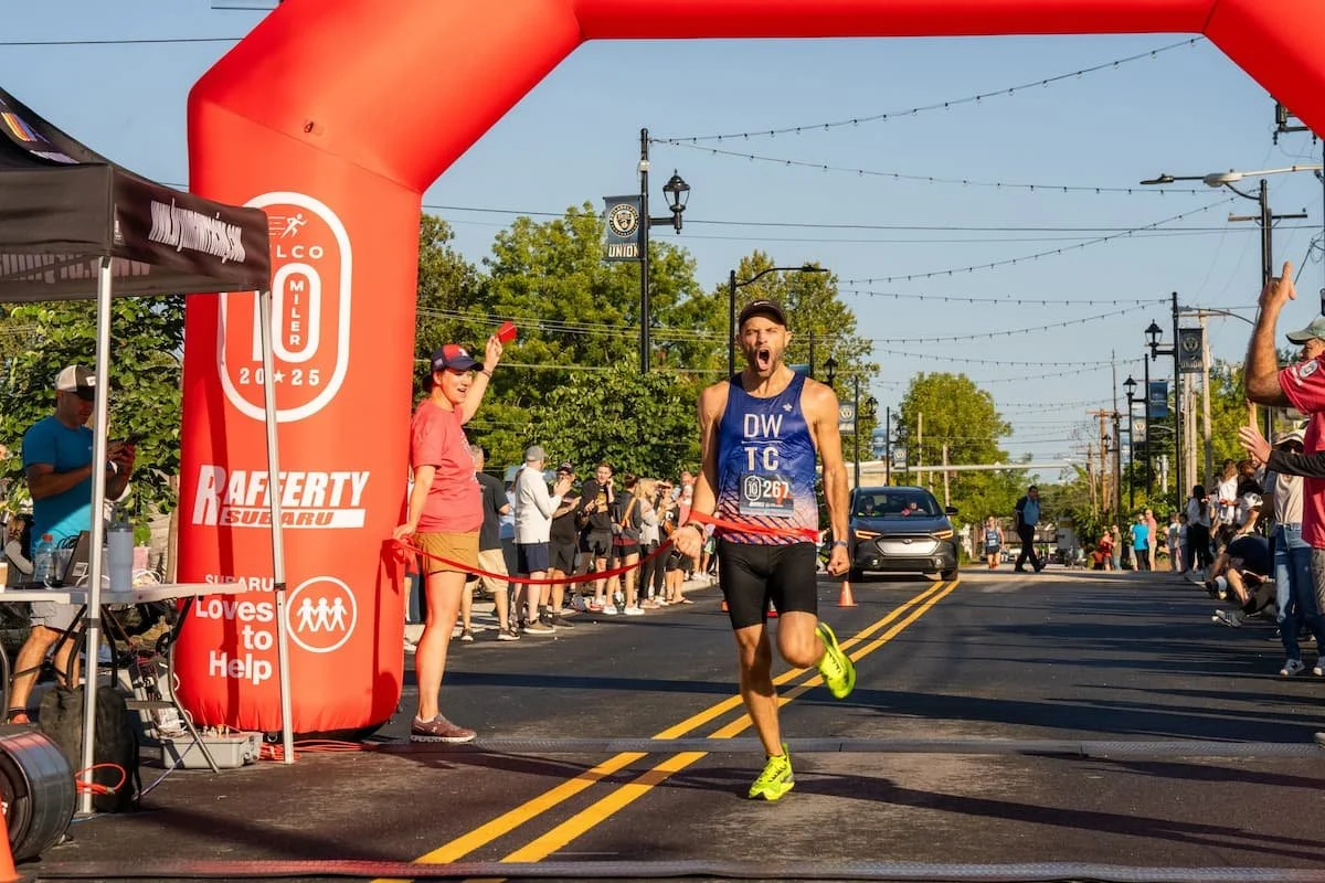 Male marathon runner crossing the finish line, celebrating, wearing a blue tank top and black shorts, in a race event with crowd and supporters in the background.