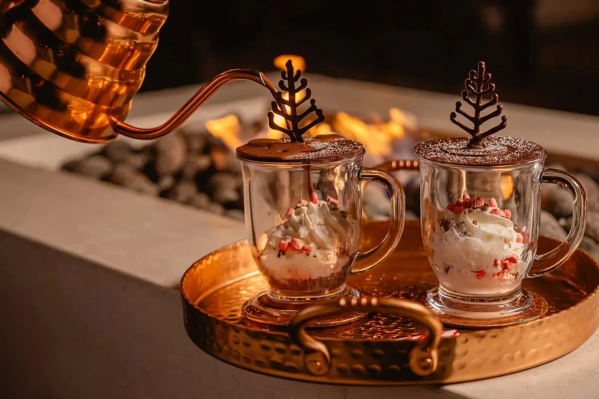 Two glass mugs of hot chocolate topped with whipped cream, red sprinkles, and chocolate decorations, sitting on a copper tray near a firepit with flames in the background.