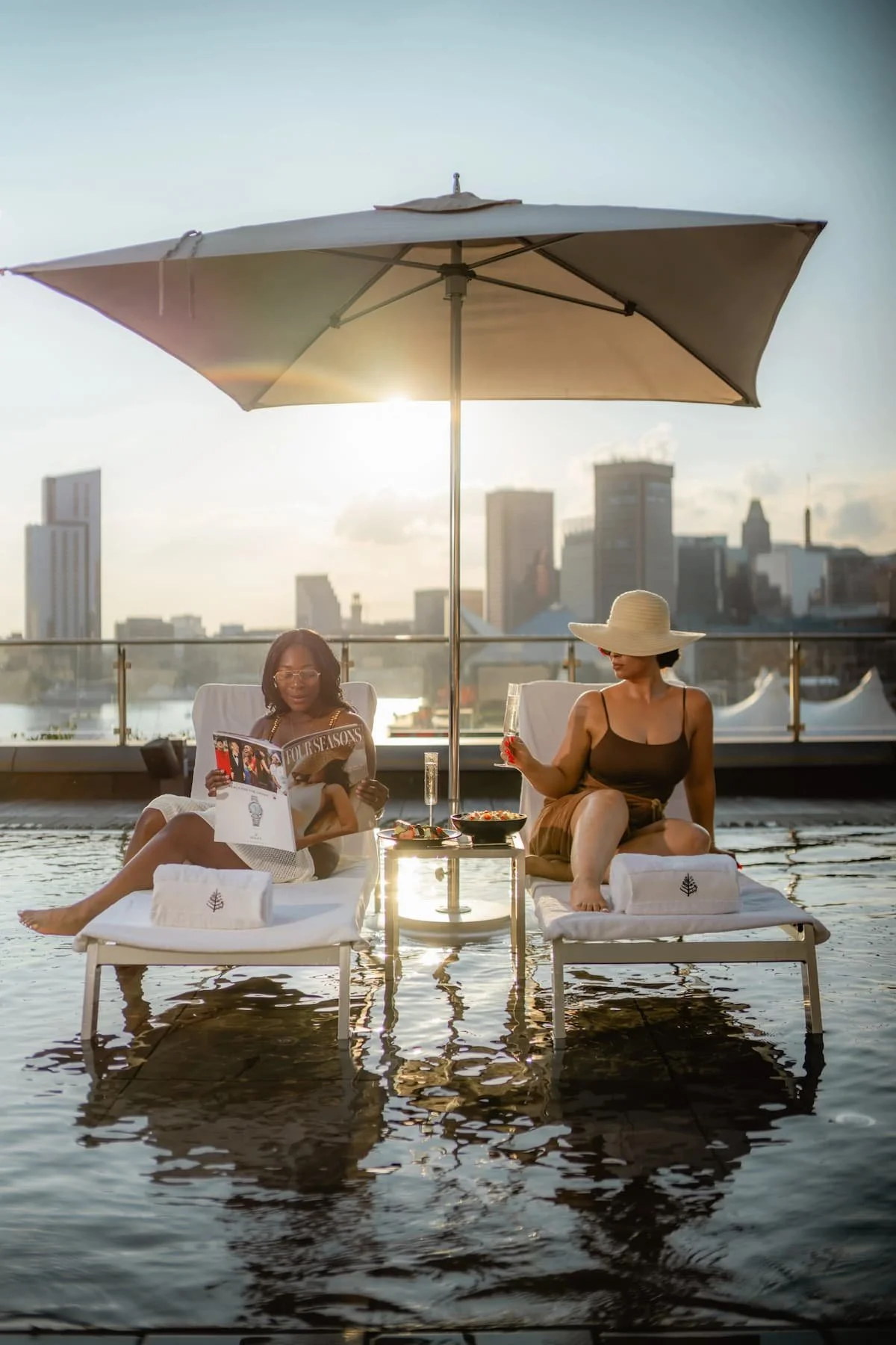 Two women relaxing on poolside lounge chairs under an umbrella with city skyline in the background, enjoying drinks and food during sunset.