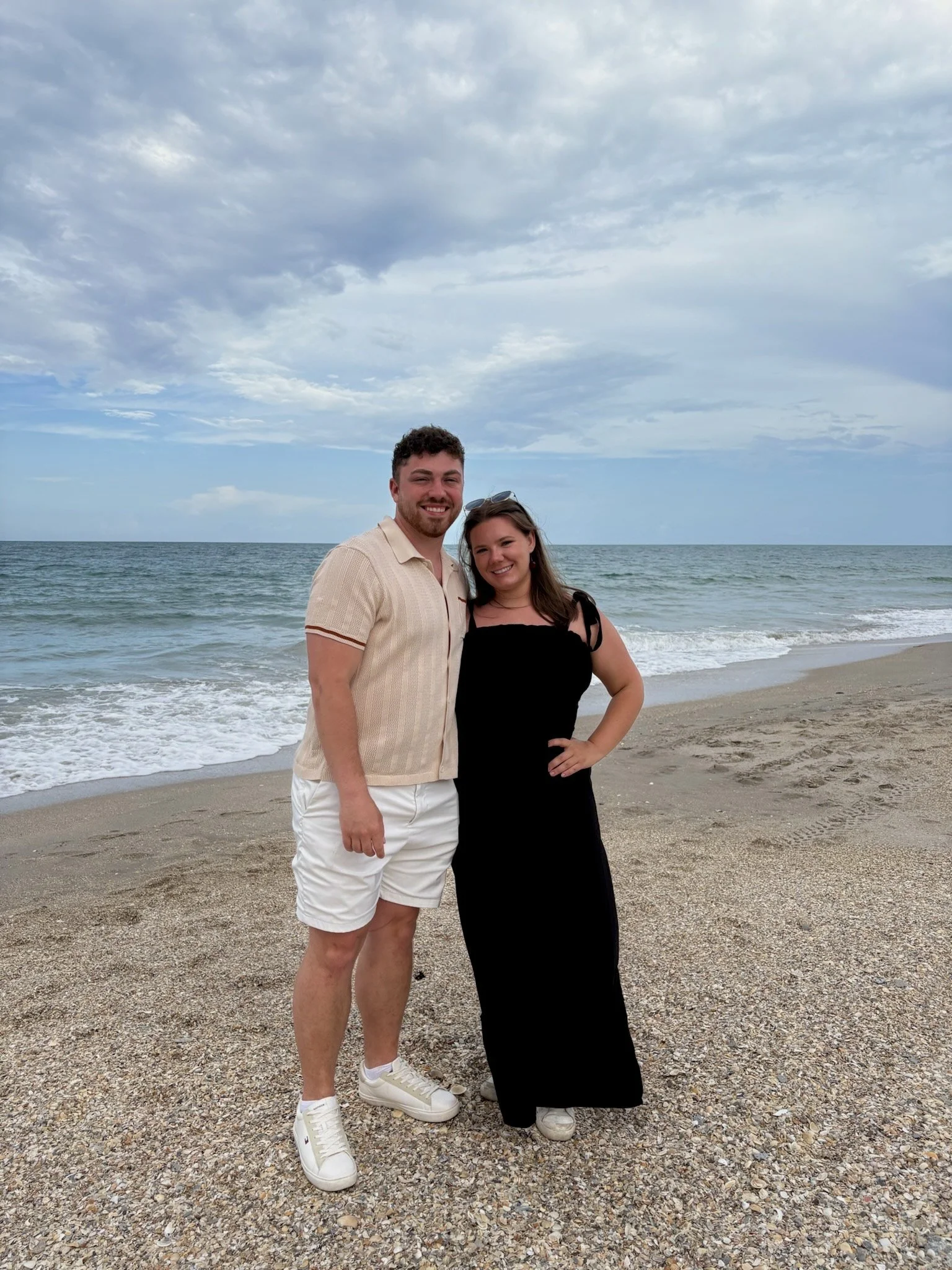 A smiling couple standing on a sandy beach near the ocean with a cloudy sky overhead.