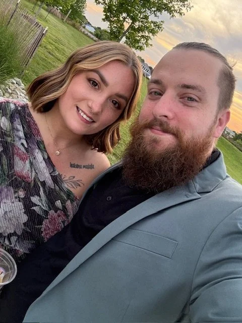 A smiling woman with shoulder-length light brown hair and a man with a beard, both dressed nicely, outdoors during sunset with green trees and a fence in the background.