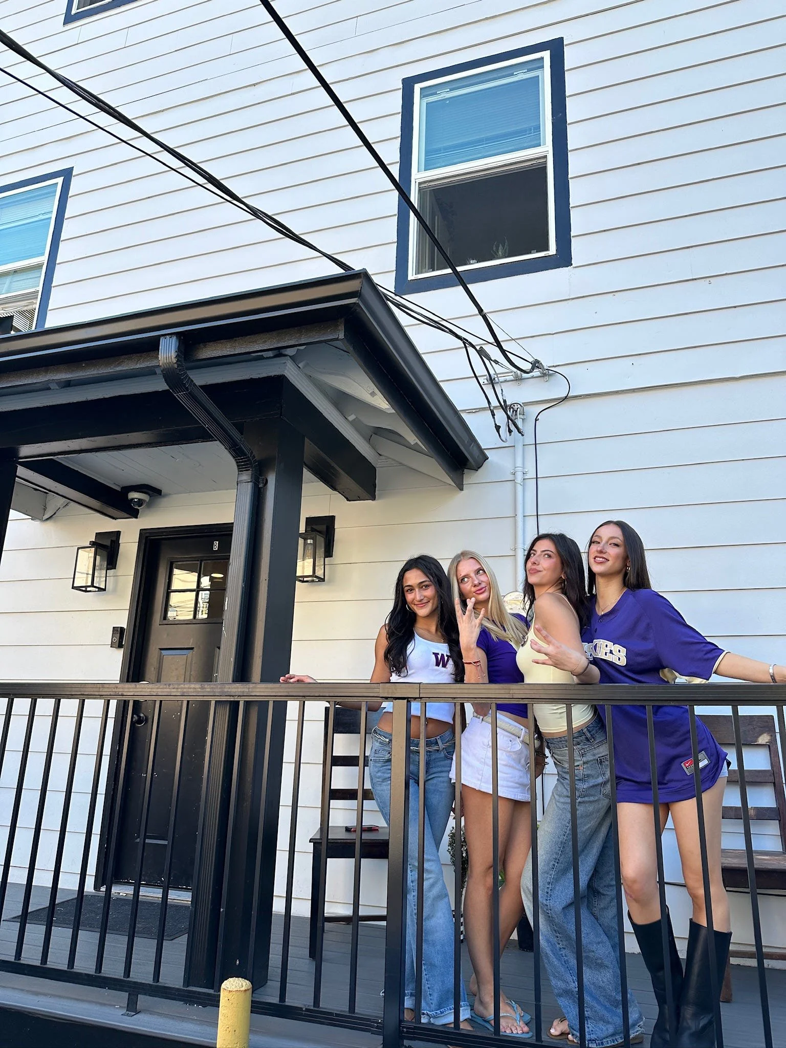 Four young women standing on a porch in front of a white house with black trim, posing and smiling for a photo.