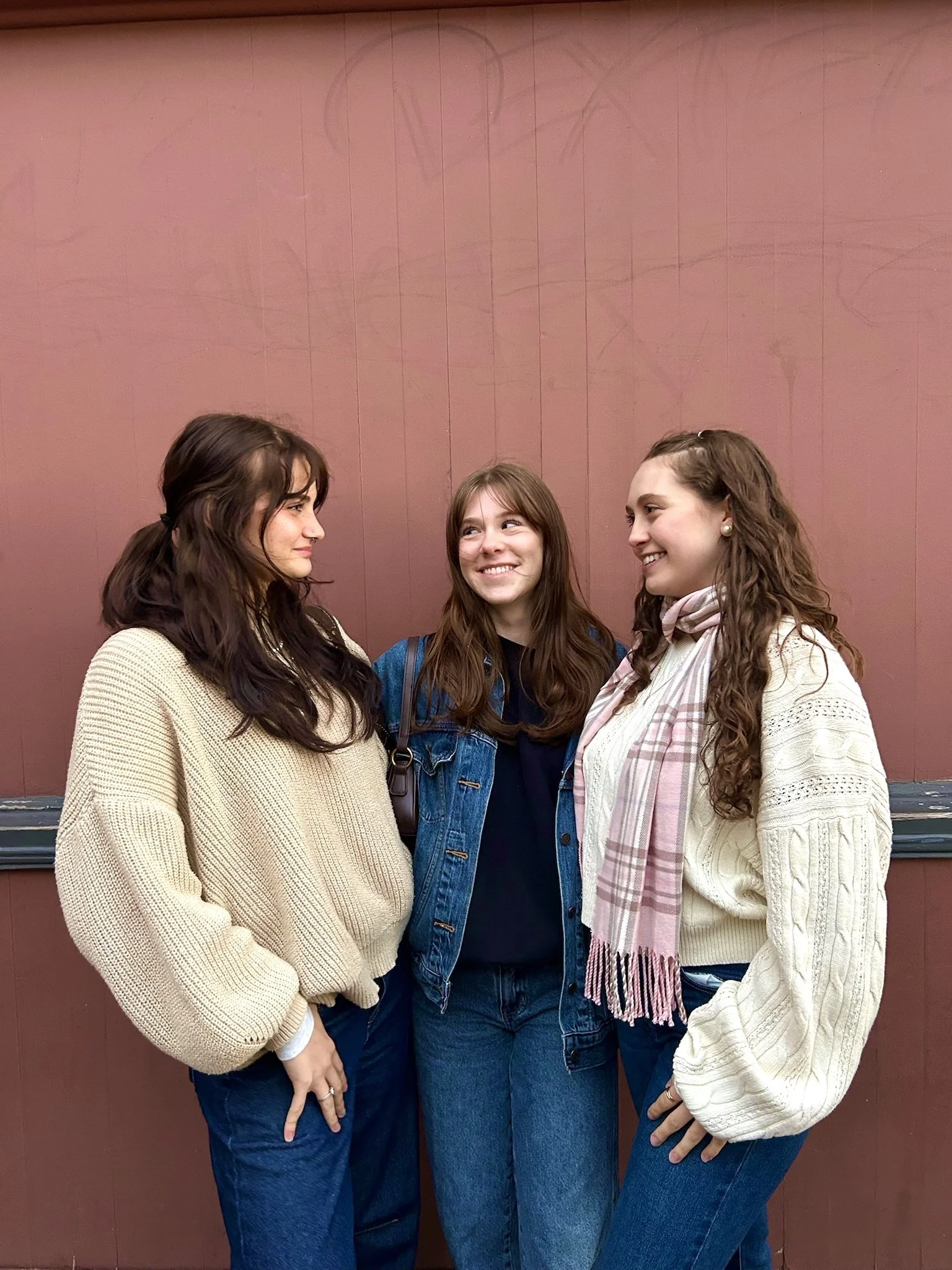 Three young women standing together in front of a pink wall, smiling and looking at each other.