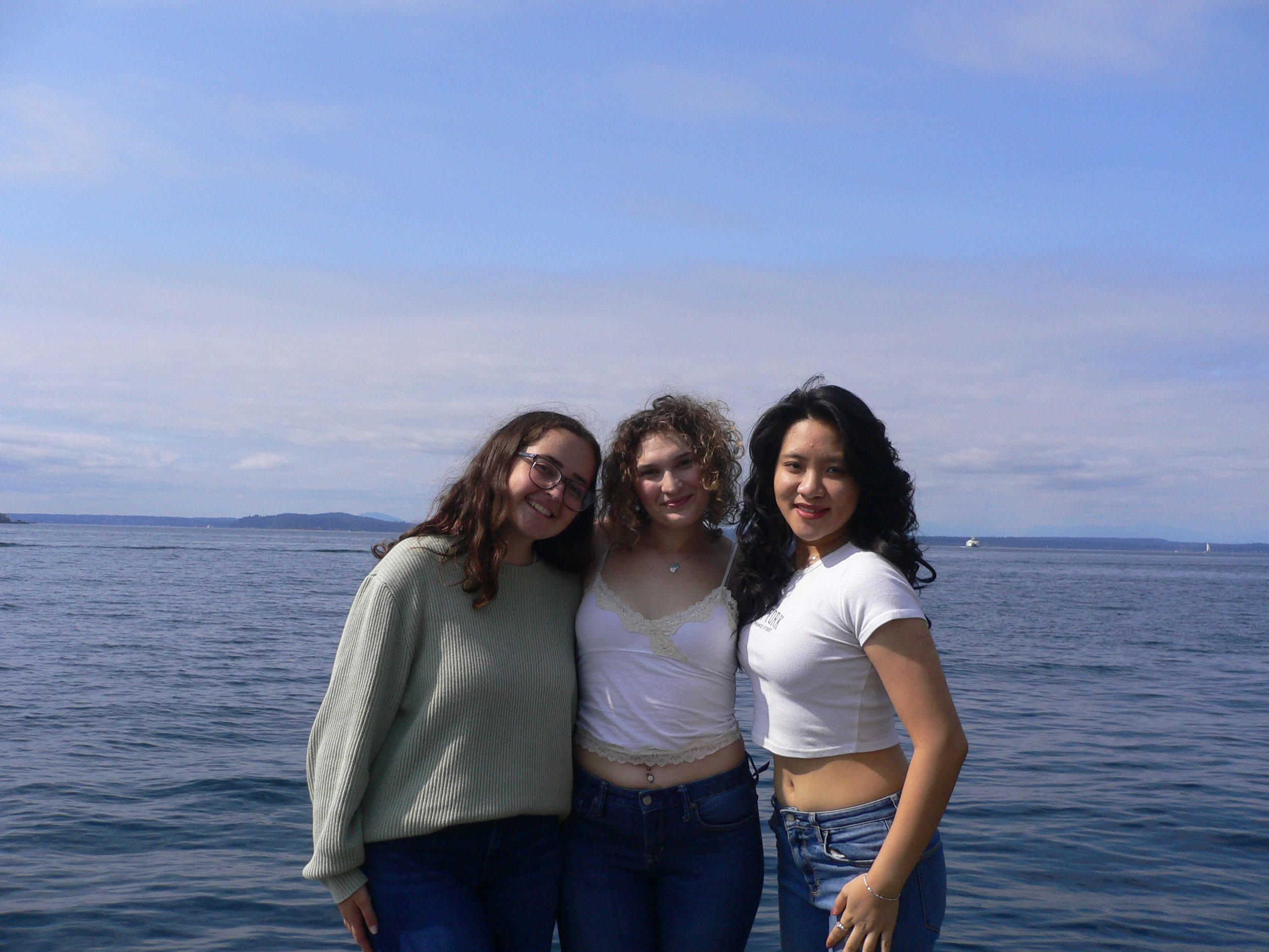 Three women standing together near a body of water with boats and a distant shoreline, smiling at the camera.