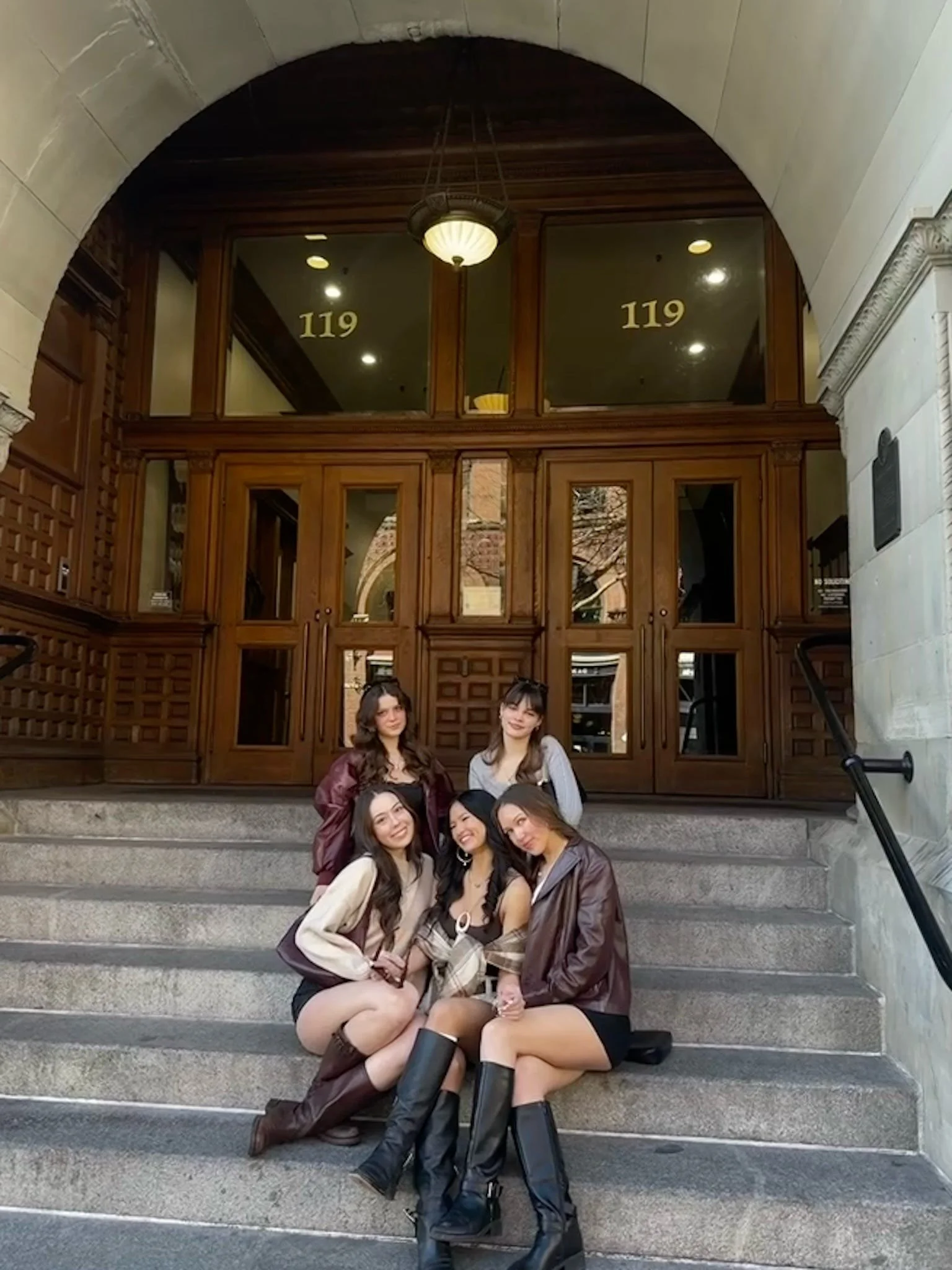 Five young women posing together on the steps outside a building with a wooden door and glass windows marked '119'.