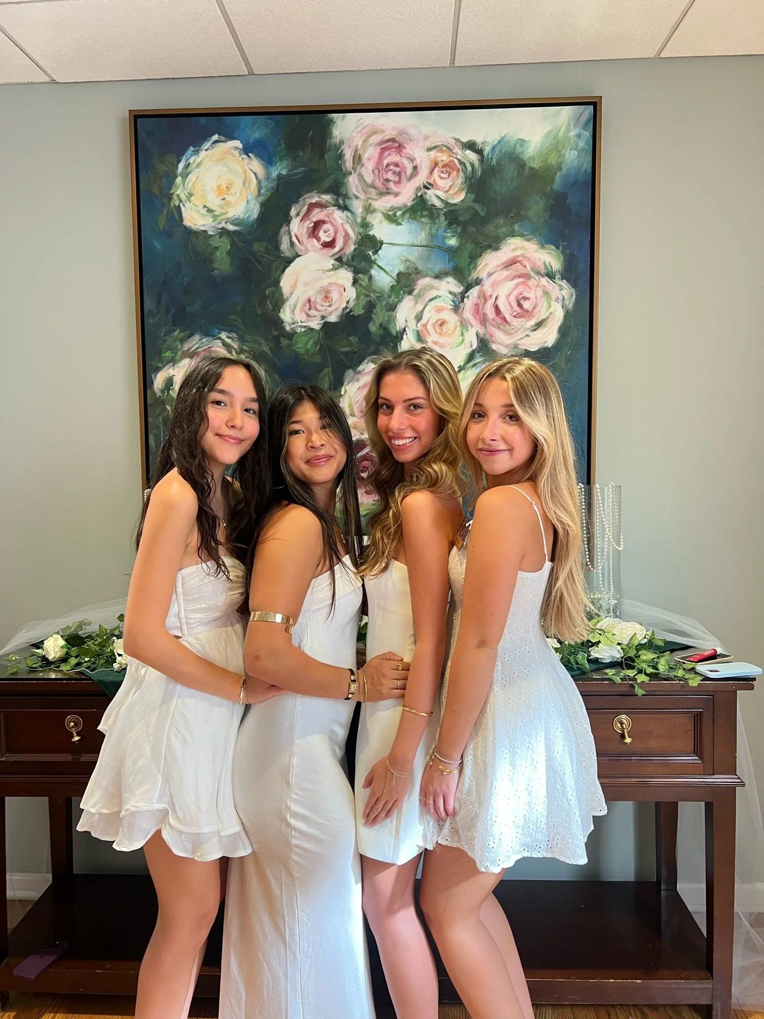 Four young women dressed in white dresses standing close together, smiling, in front of a floral painting and a table with white roses and jewelry.