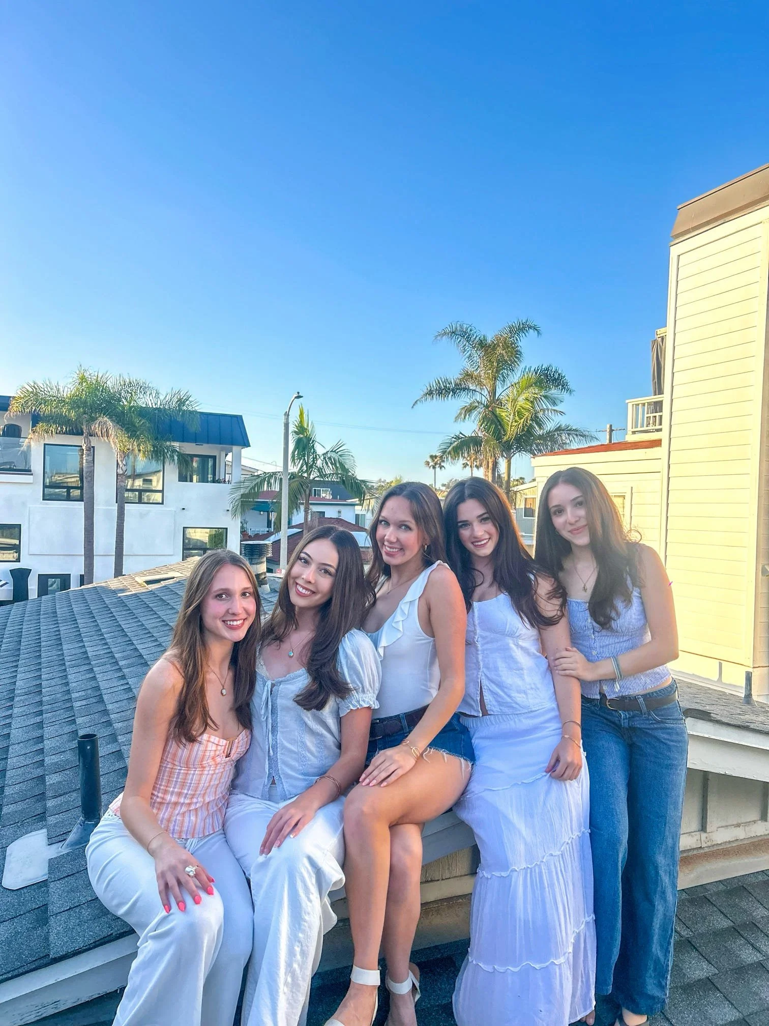 Five young women sitting on a rooftop during daytime with palm trees and buildings in the background, all smiling.