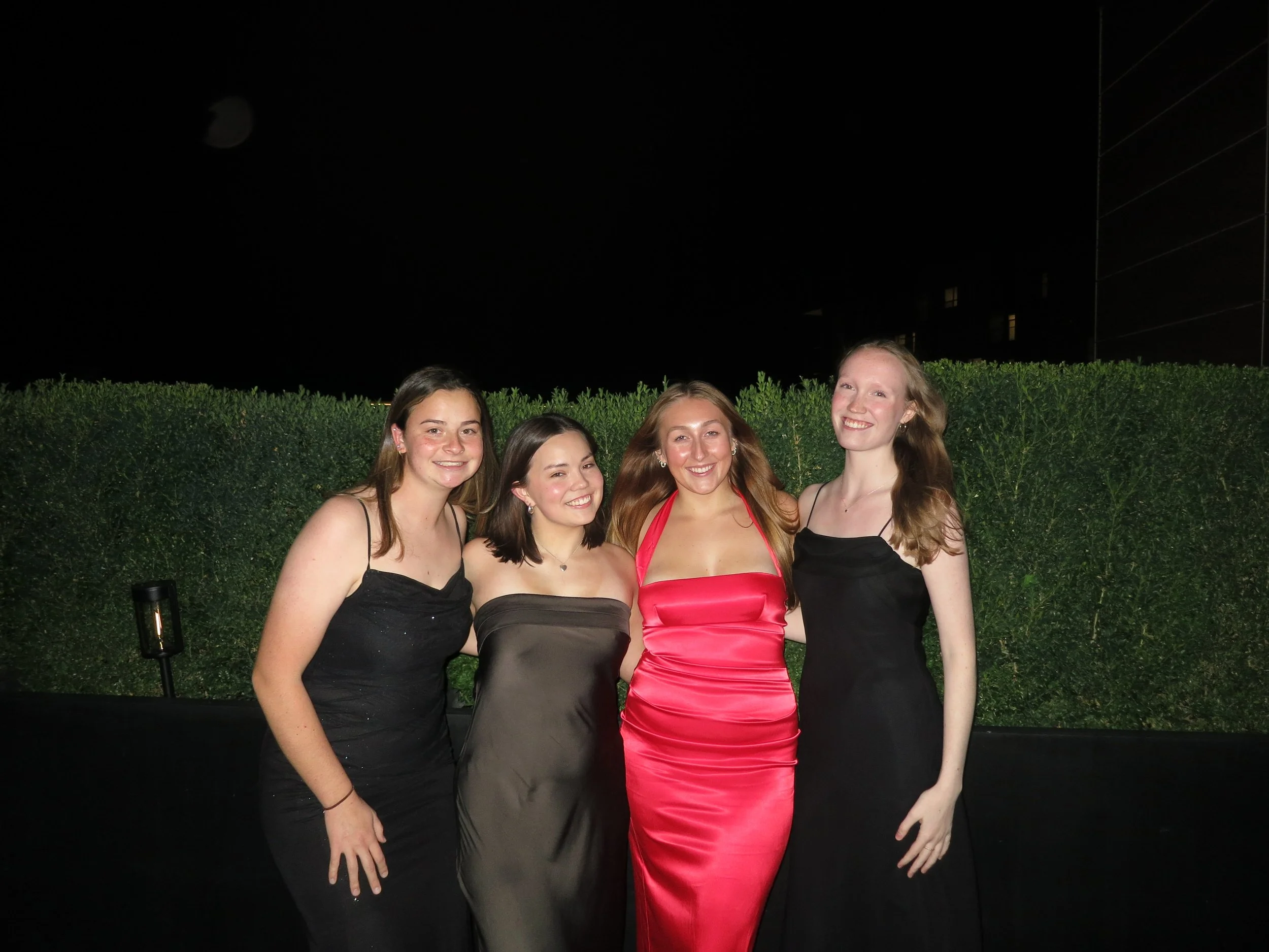 Four young women in elegant dresses, smiling and posing outdoors at night near a green hedge.