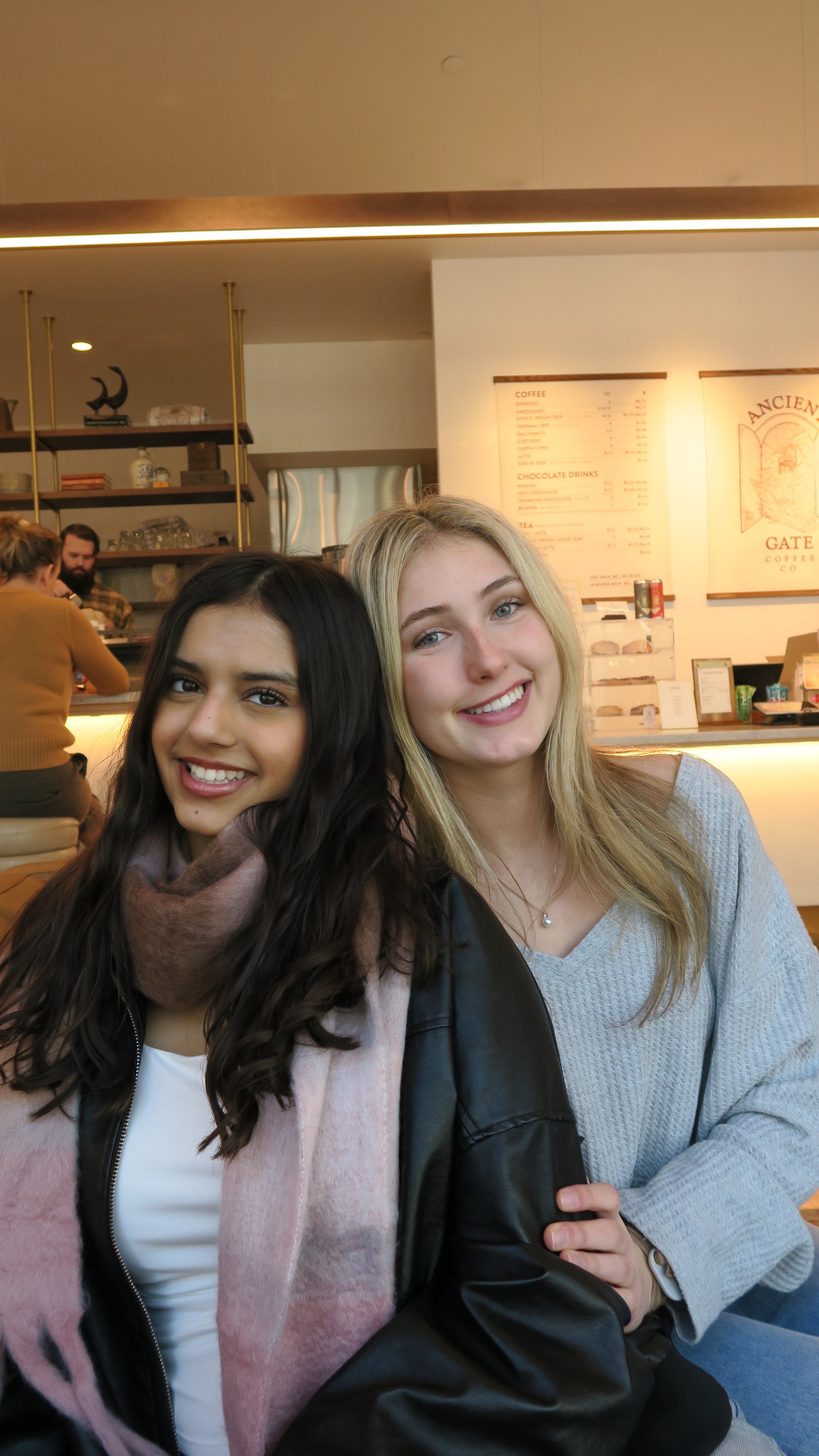 Two smiling women sitting at a cafe counter, with the cafe menu and staff visible in the background.