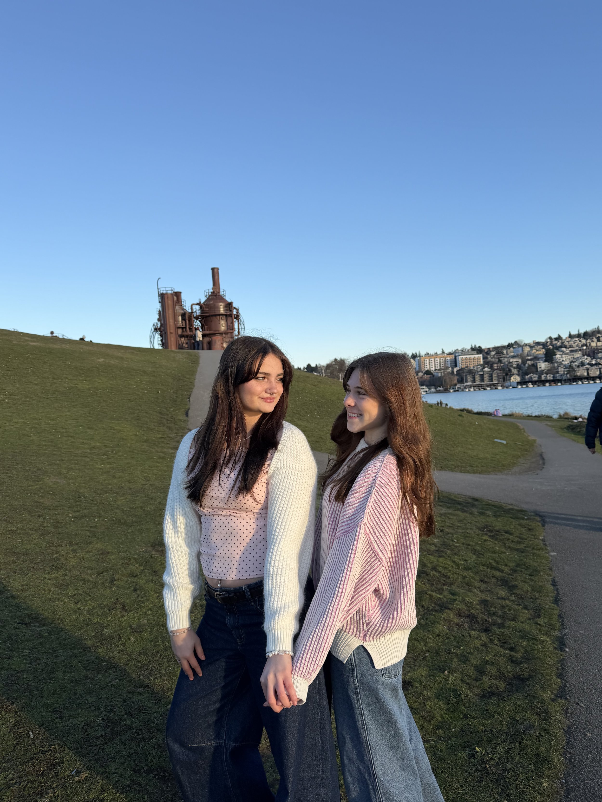 Two young women holding hands outdoors near a body of water during sunset, with industrial structures and buildings in the background.