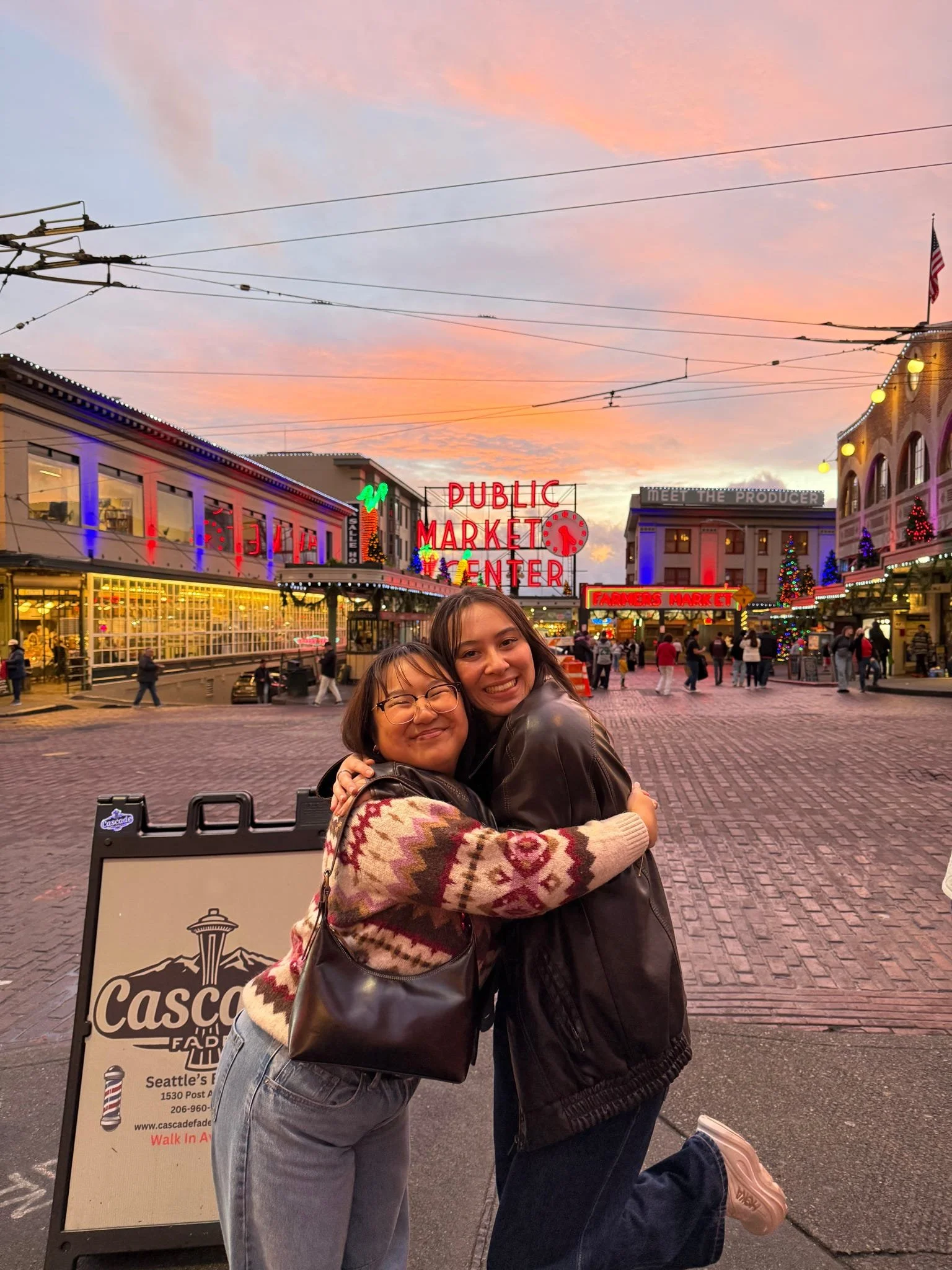 Two women hugging and smiling in front of Pike Place Market at sunset, with neon signs and decorated buildings in Seattle, Washington.