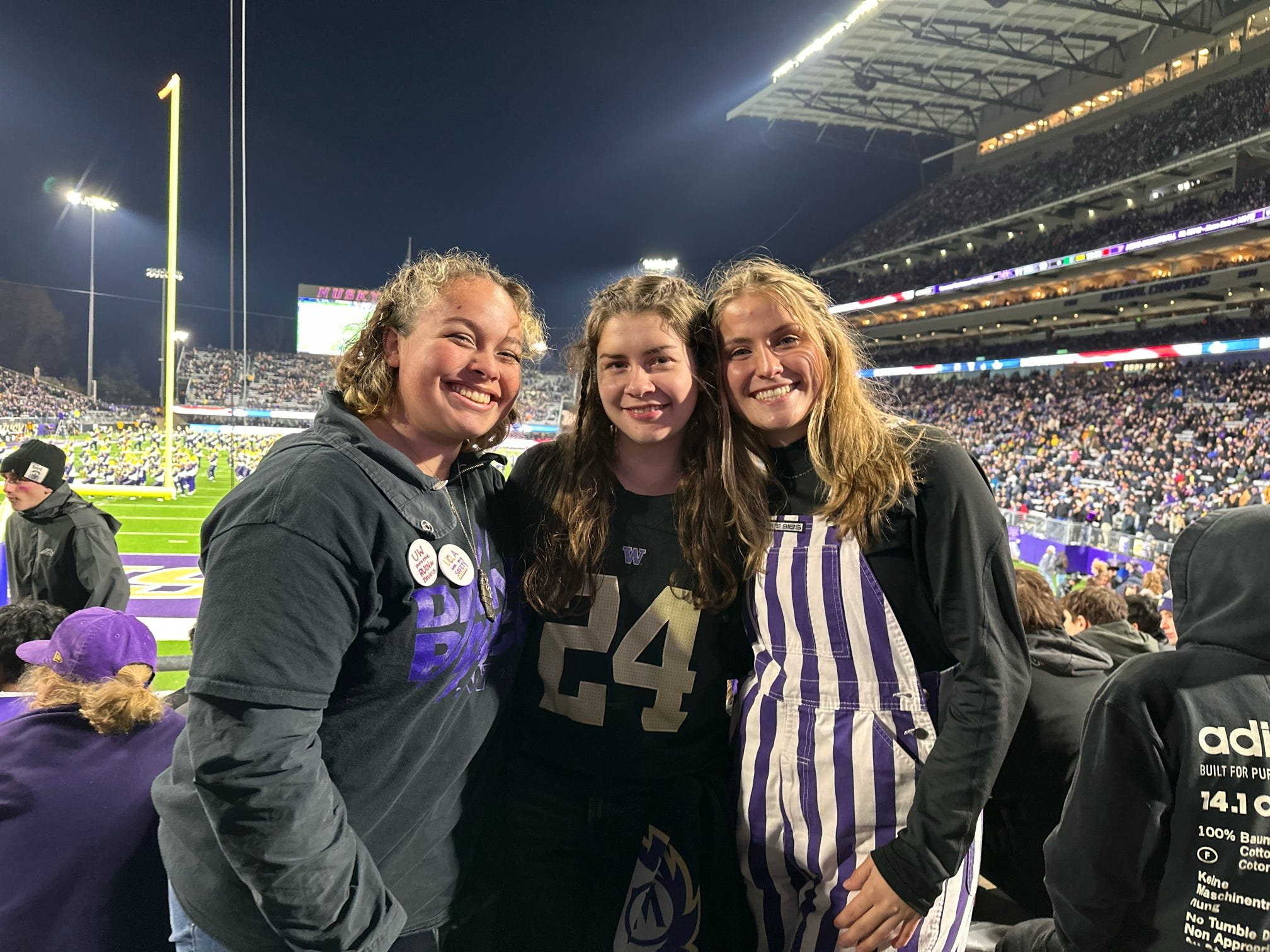 Three young women smiling at a football game, standing in the stadium near the field with a large crowd behind them on a night game.