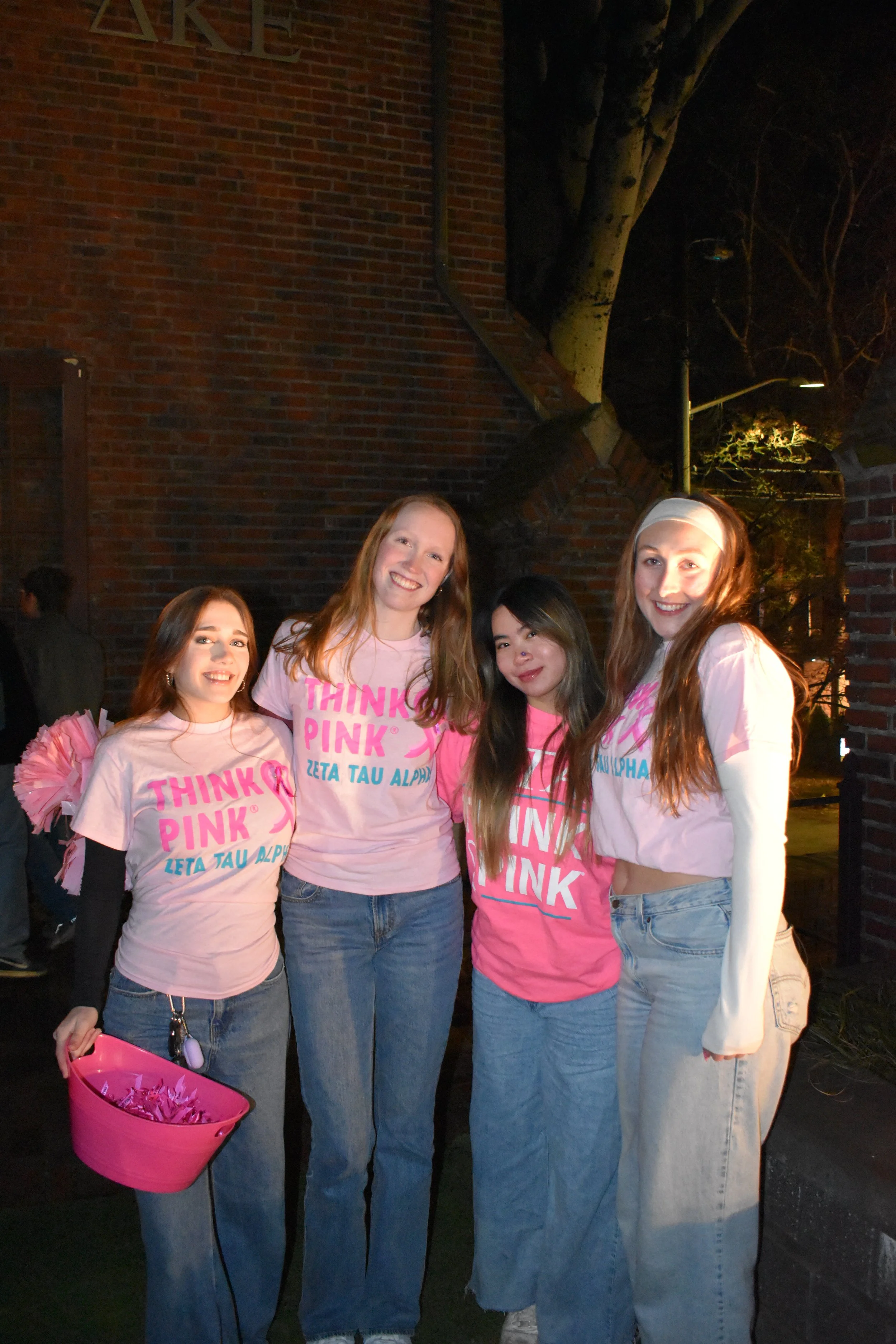Four young women stand together outdoors at night, wearing pink T-shirts with the words 'Think Pink' and Greek letters, smiling at the camera.