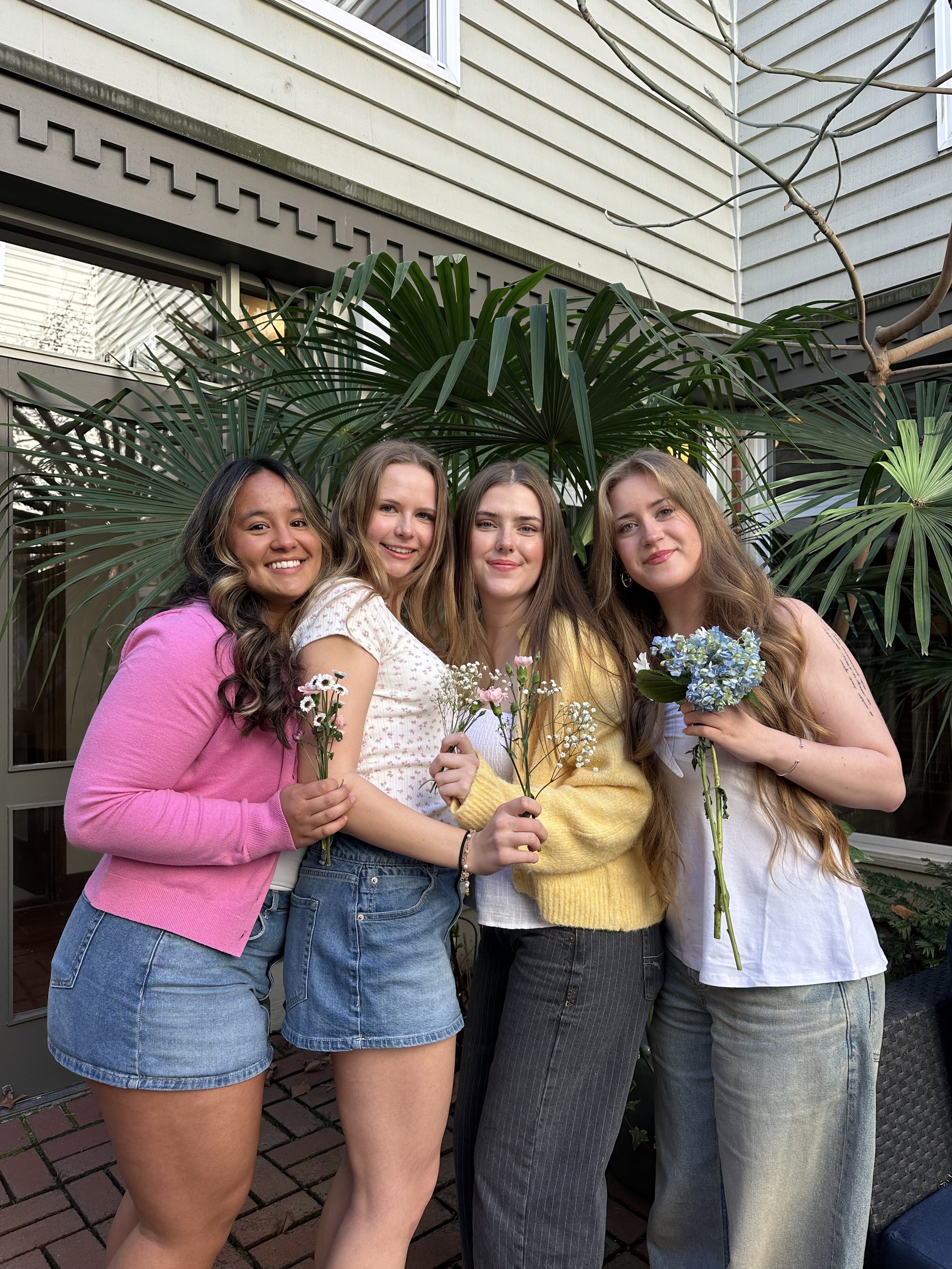 Four young women smiling and holding flowers outdoors in front of large green plants, in a backyard or patio area.