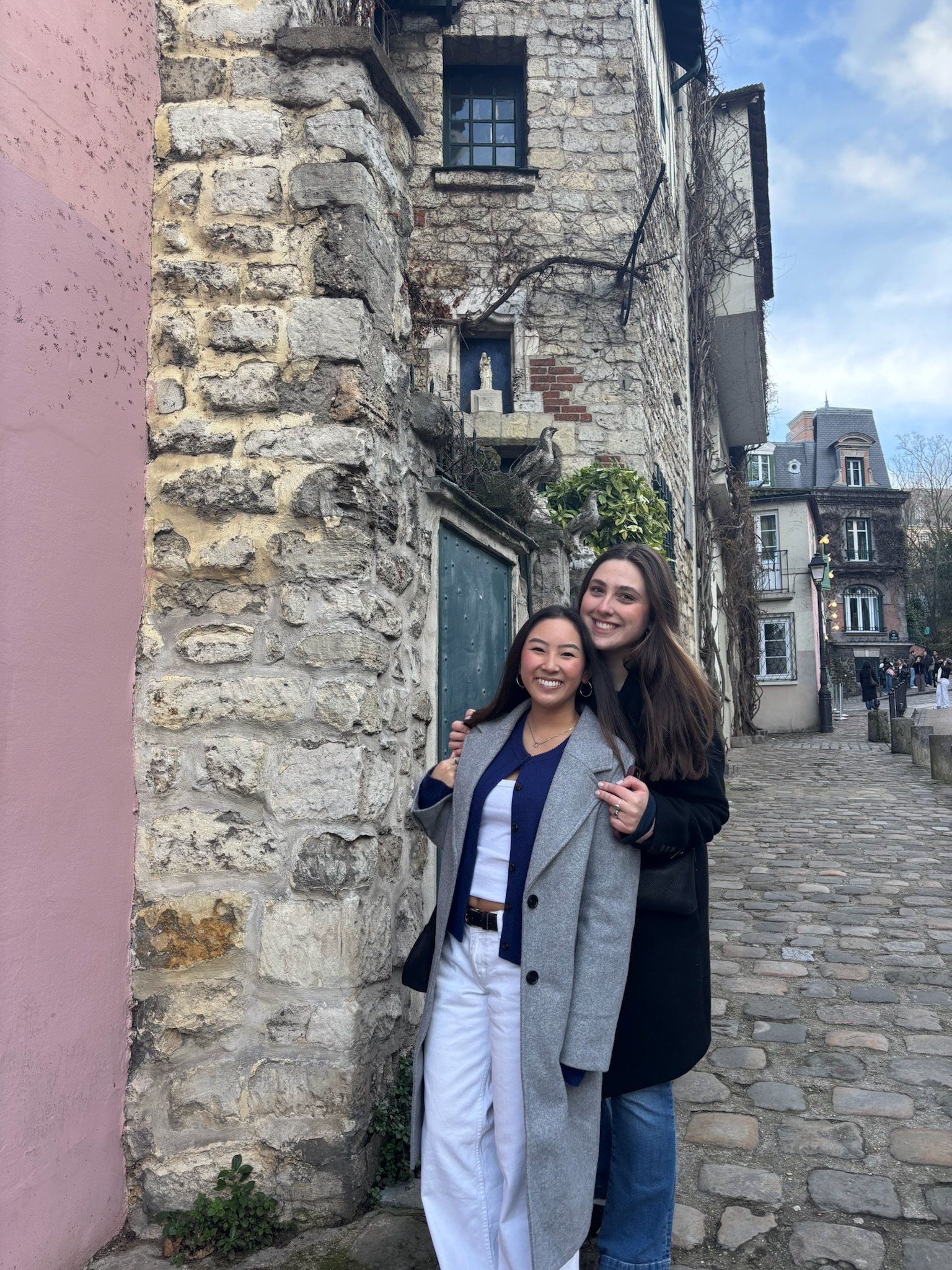 Two women smiling and hugging on a cobblestone street in front of a stone and brick building with decorative elements. One woman wears a gray coat and white pants, and the other wears a black coat and blue jeans.