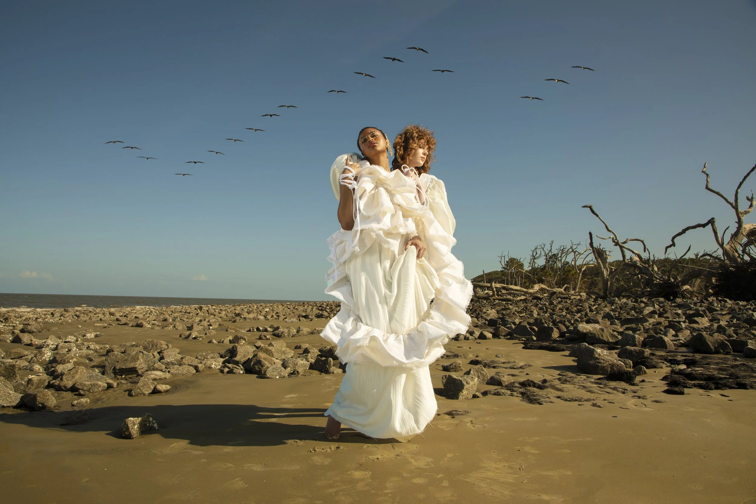 Two women in white dresses standing on a rocky beach with a clear blue sky, flying seagulls, and driftwood in the background.