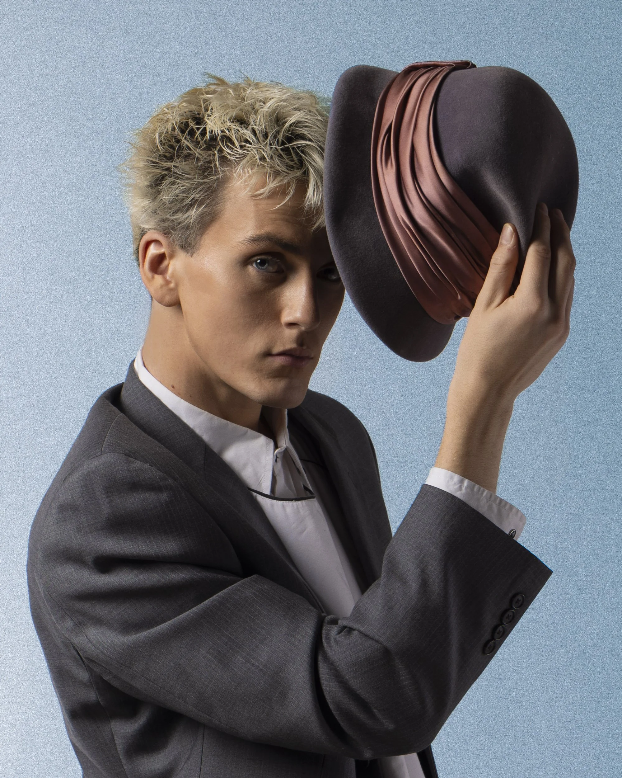 A person with short, curly blonde hair, wearing a dark gray suit, and holding a fashionable dark brown hat with a decorative reddish-brown band, partially obscures part of their face against a light blue background.