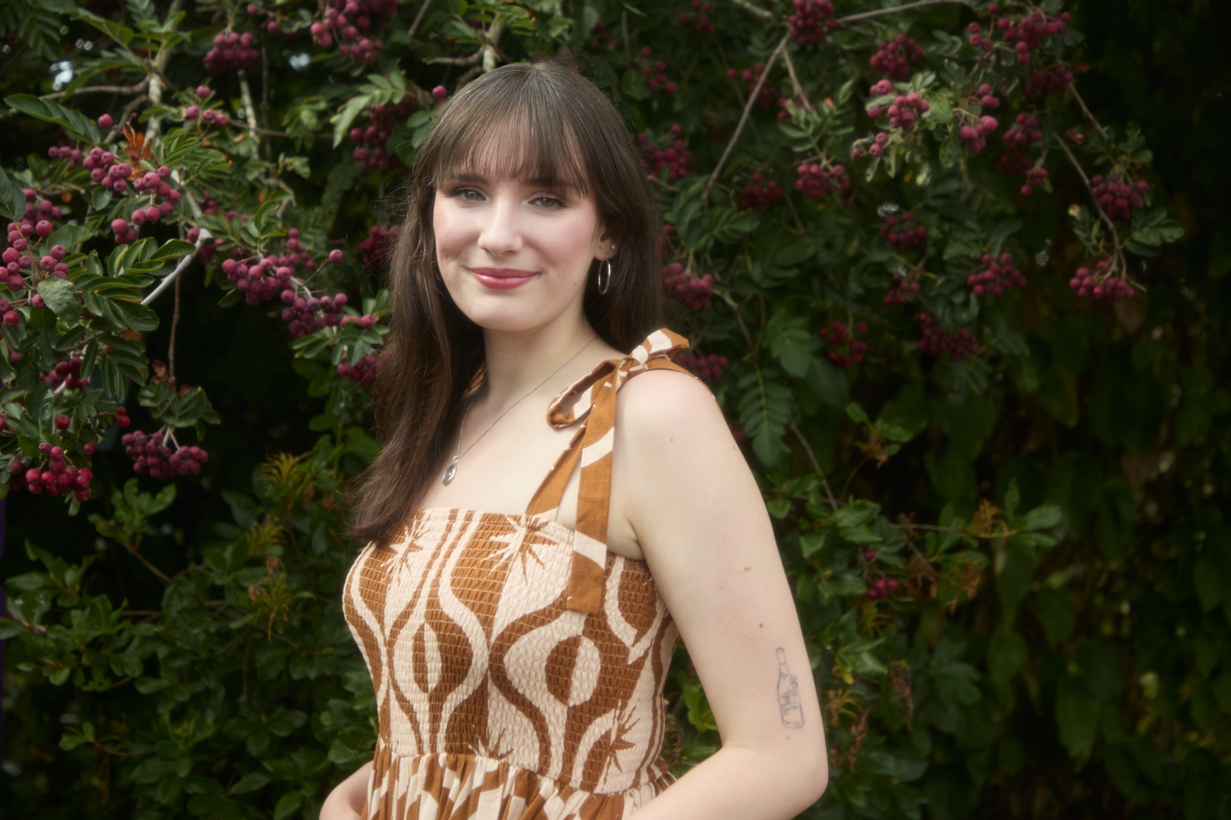 A young woman with brown hair and bangs smiling outdoors in front of a bush with dark green leaves and clusters of small purple berries. She is wearing a sleeveless dress with a brown and beige pattern, a silver necklace with a pendant, and hoop earr
