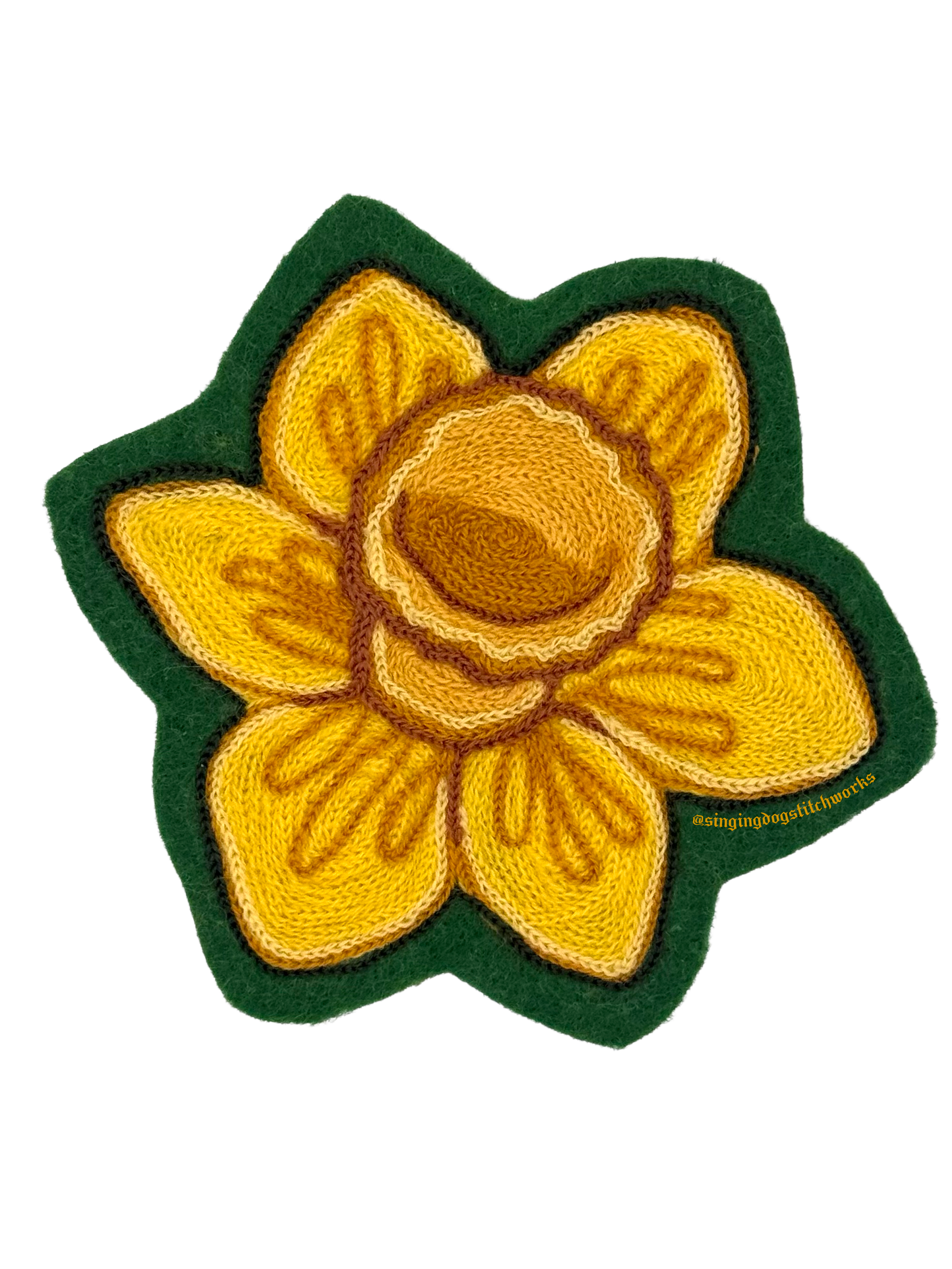 A close-up of a yellow flower-shaped embroidered patch with brown accents, with a green border and backing, and the text '@singingdogstitchworks' in yellow at the bottom right.