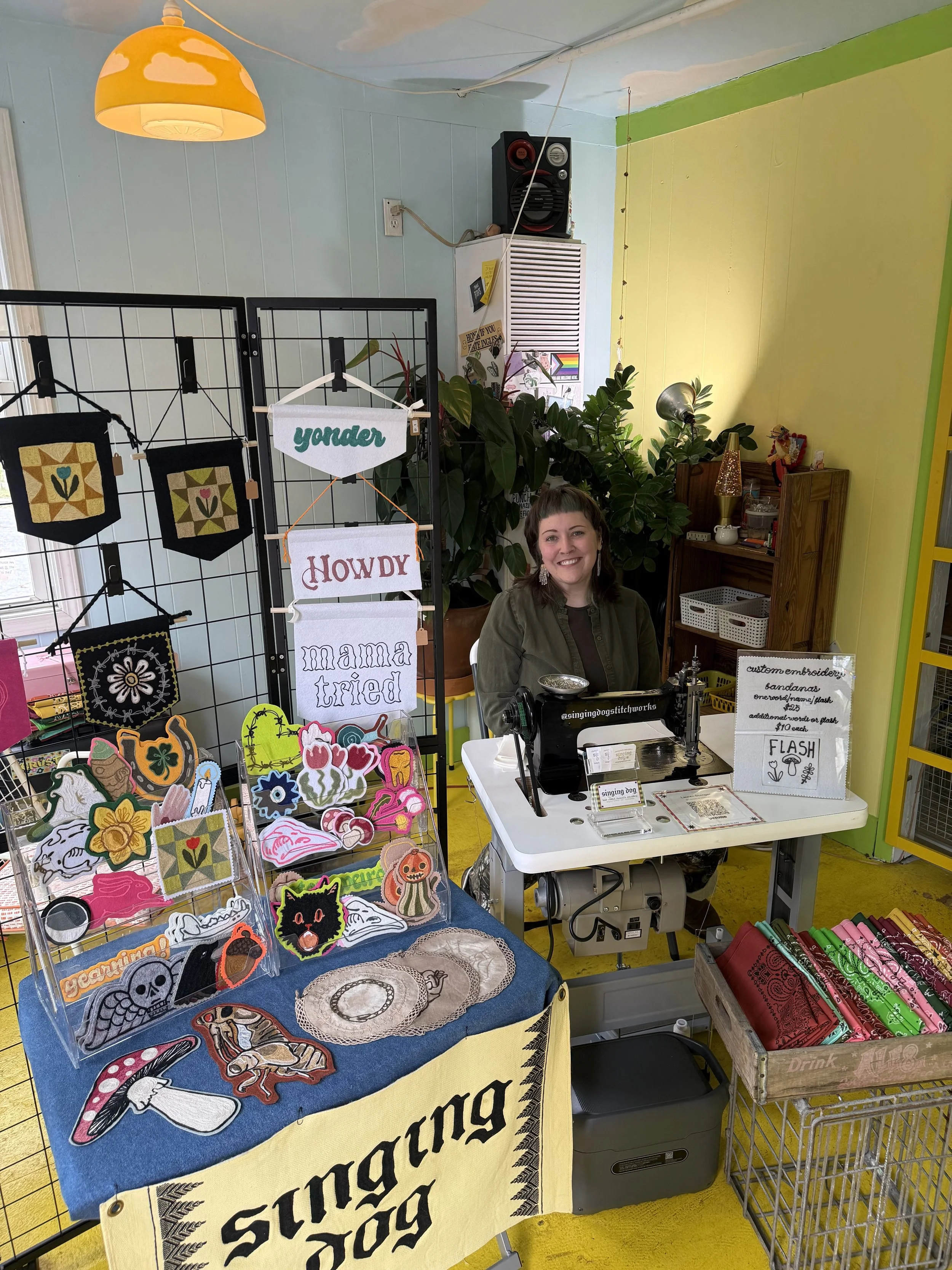 A woman sitting at a craft booth with embroidered patches, banners, and embroidered items on display. The booth has a sewing machine and a sign advertising custom embroidery.