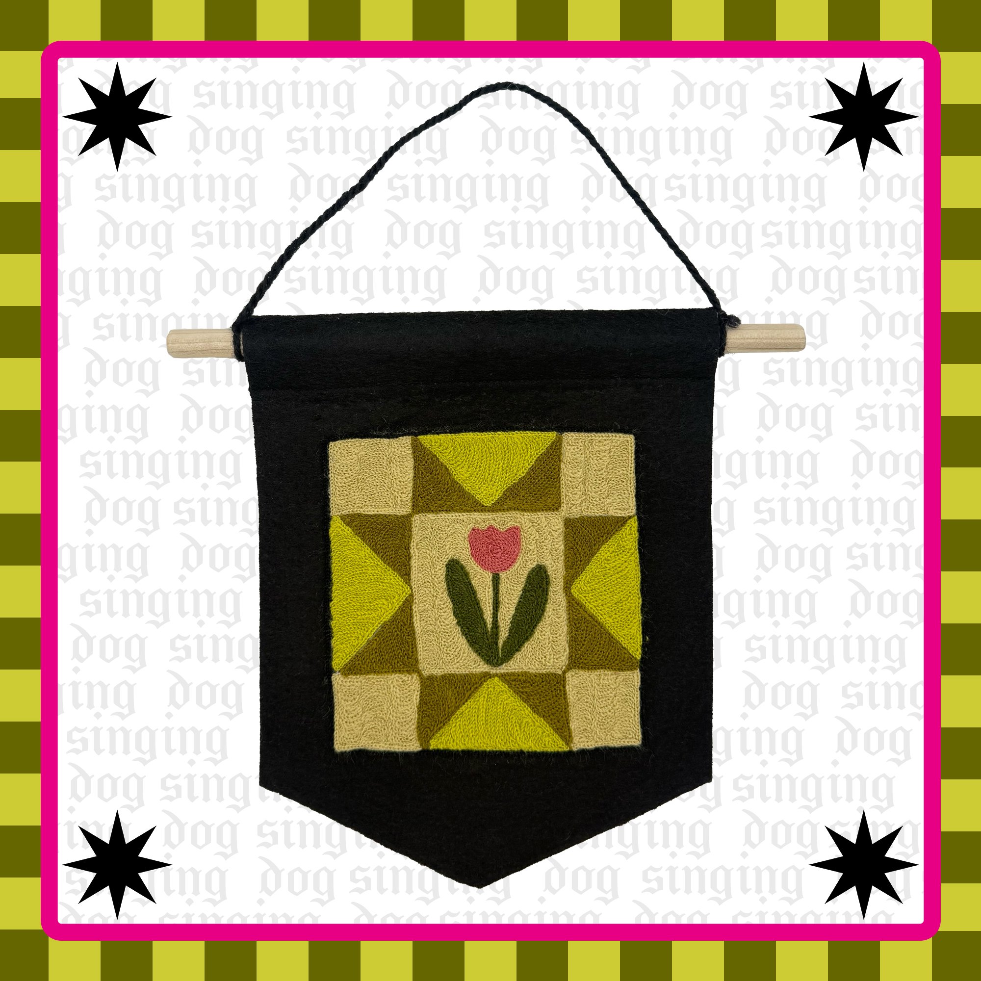 A handmade wall hanging featuring a geometric pattern with a tulip design at the center, attached to a black felt back with a wooden dowel and hanging cord, decorative stars in each corner, on a white background bordered by a pink and green frame.