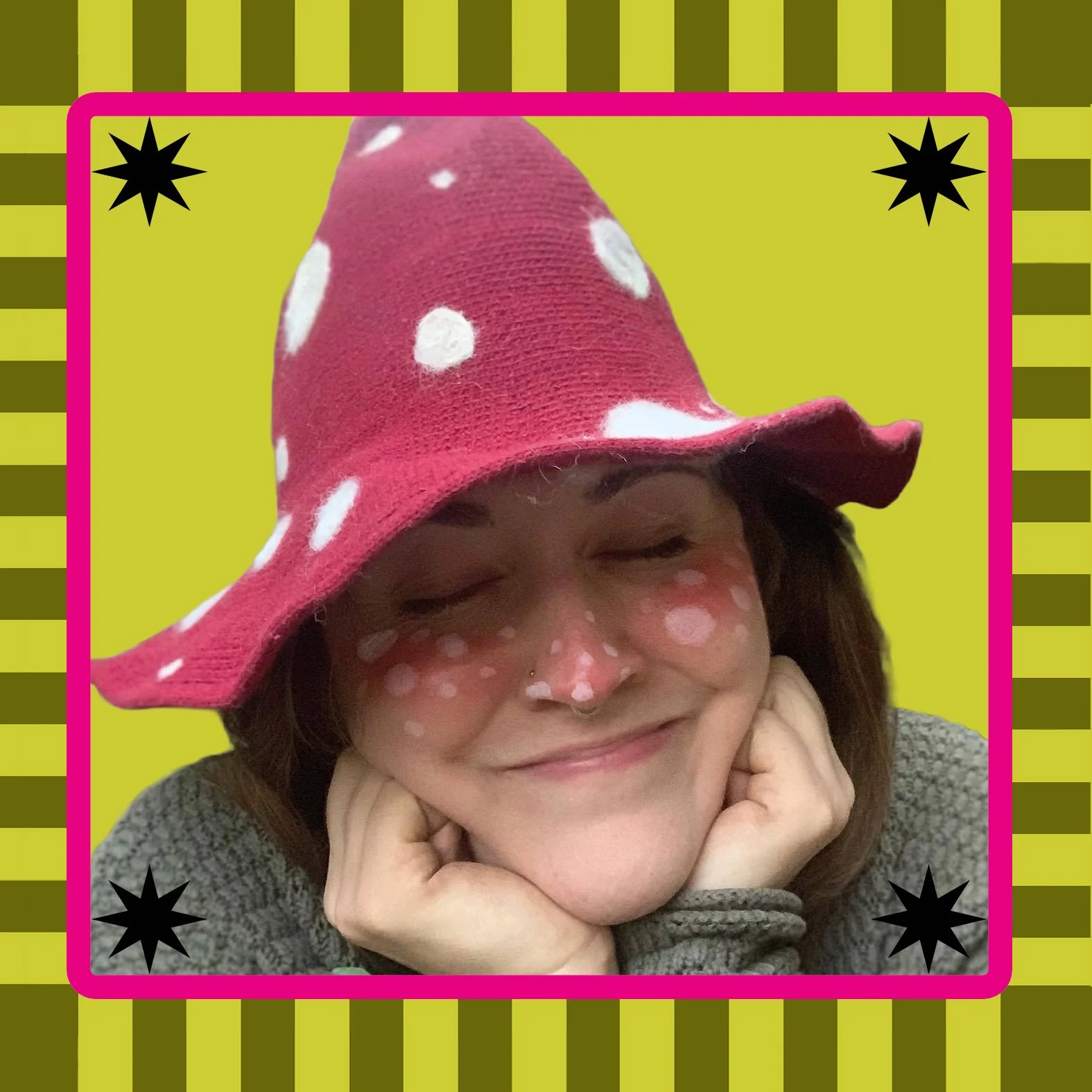 Cute picture of a girl in a green sweater wearing a red mushroom cap as a hat with her cheeks covered in blush and white spots.