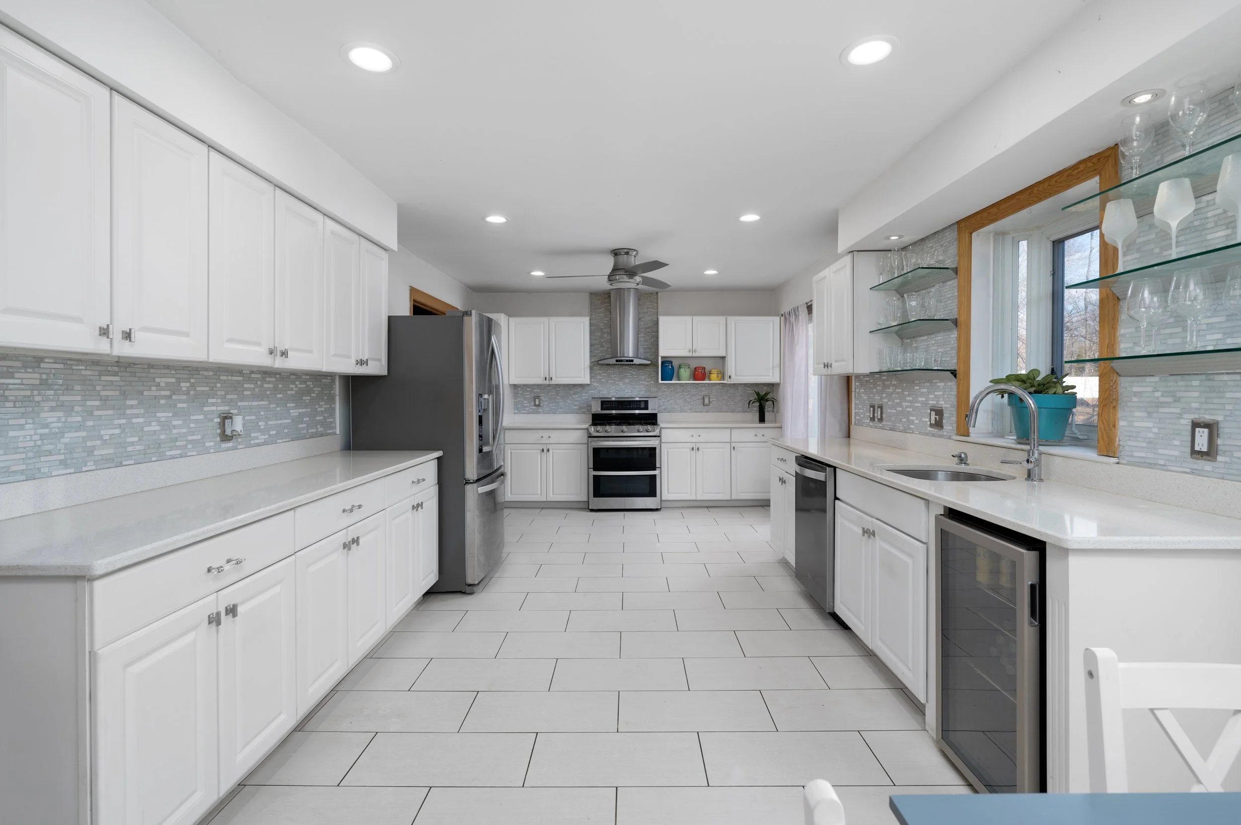 Modern white kitchen with gray backsplash, stainless steel appliances, open shelving, and a large window with a blue plant pot.