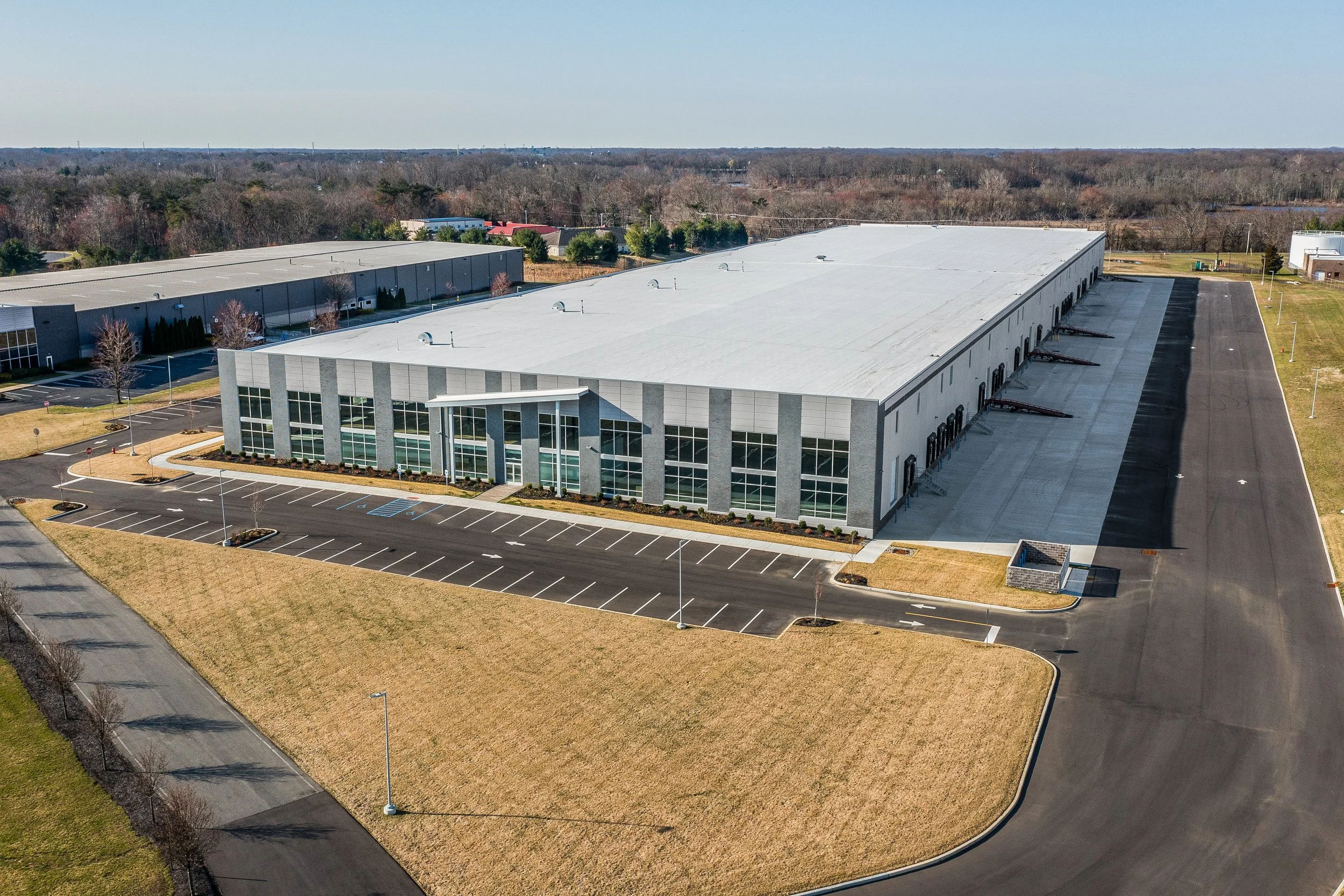 A large, modern industrial or warehouse building with a white roof, glass entrance, and striped gray facade, surrounded by an empty parking lot and green grass, located in a suburban area.