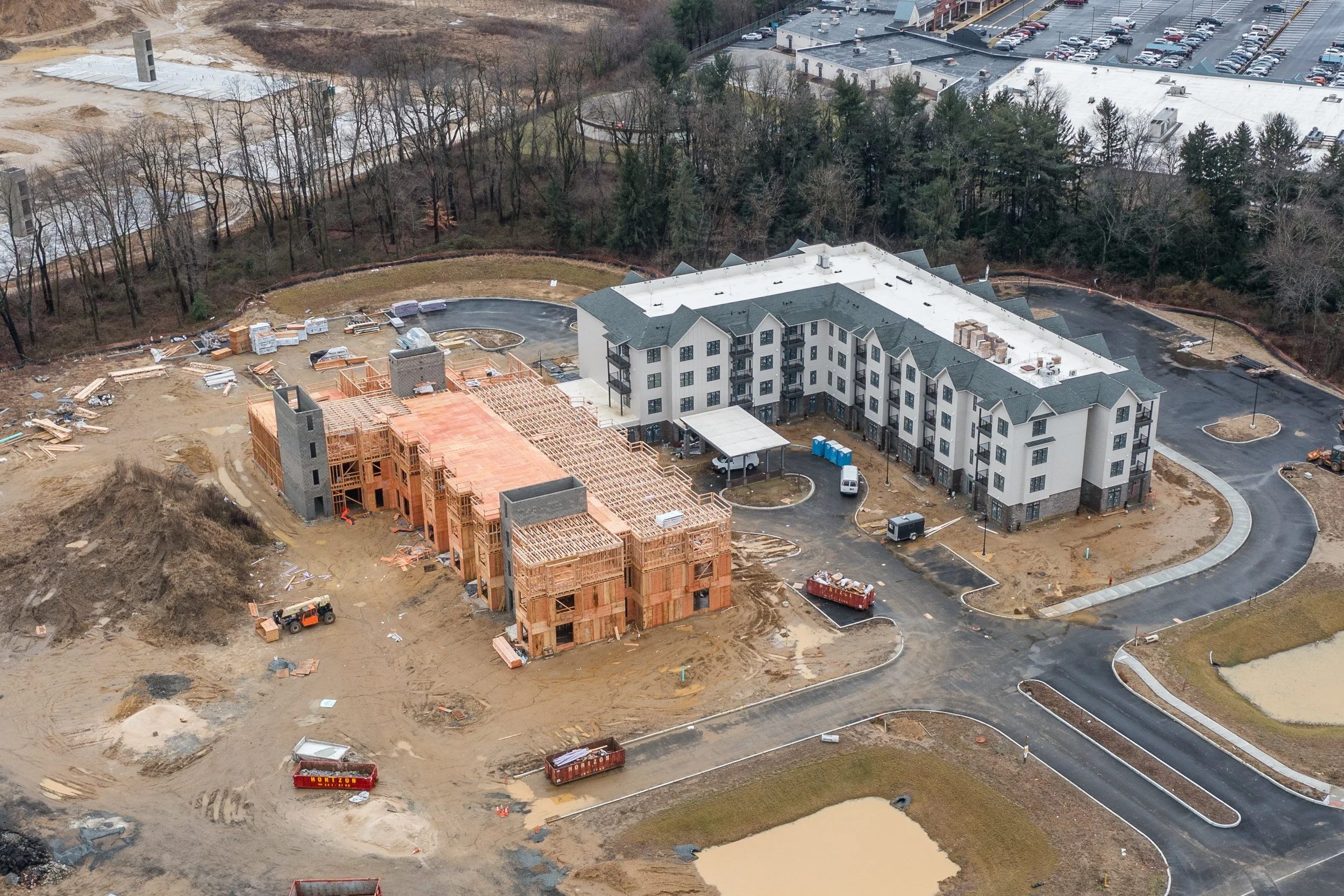 Aerial view of a construction site with a mix of unfinished wooden structures and a nearly completed multi-story white building with dark roofing, surrounded by parking lots, roads, trees, and a small pond.