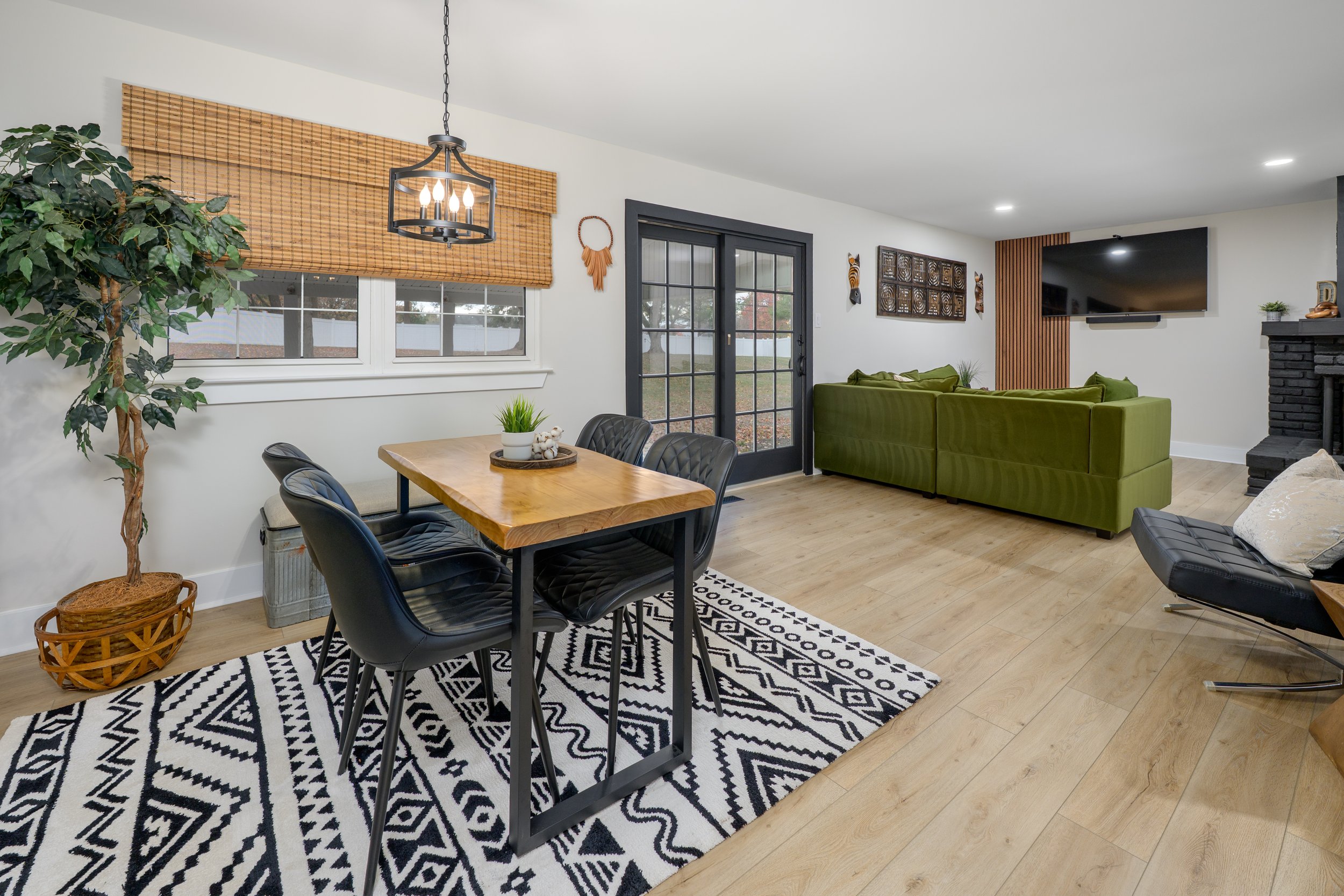 A living room and dining area with a black and white patterned rug, a wooden dining table with four black chairs, a large green sectional sofa, a wall-mounted TV, decorative wall art, and a large potted plant near a window with a woven bamboo shade.