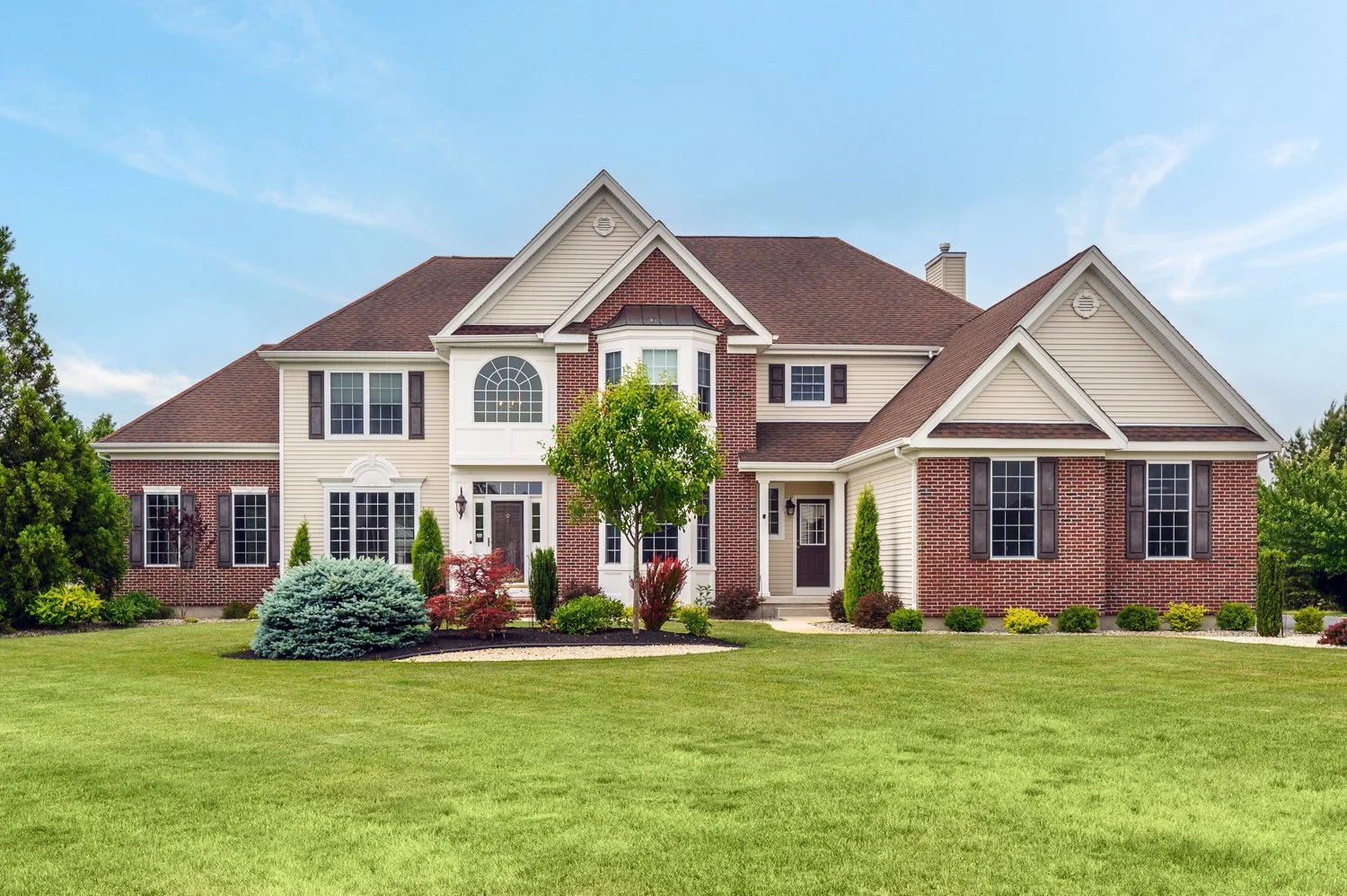 Large, two-story house with brick and siding exterior, multiple gabled roofs, and a well-kept front yard with green grass, shrubs, and small trees.