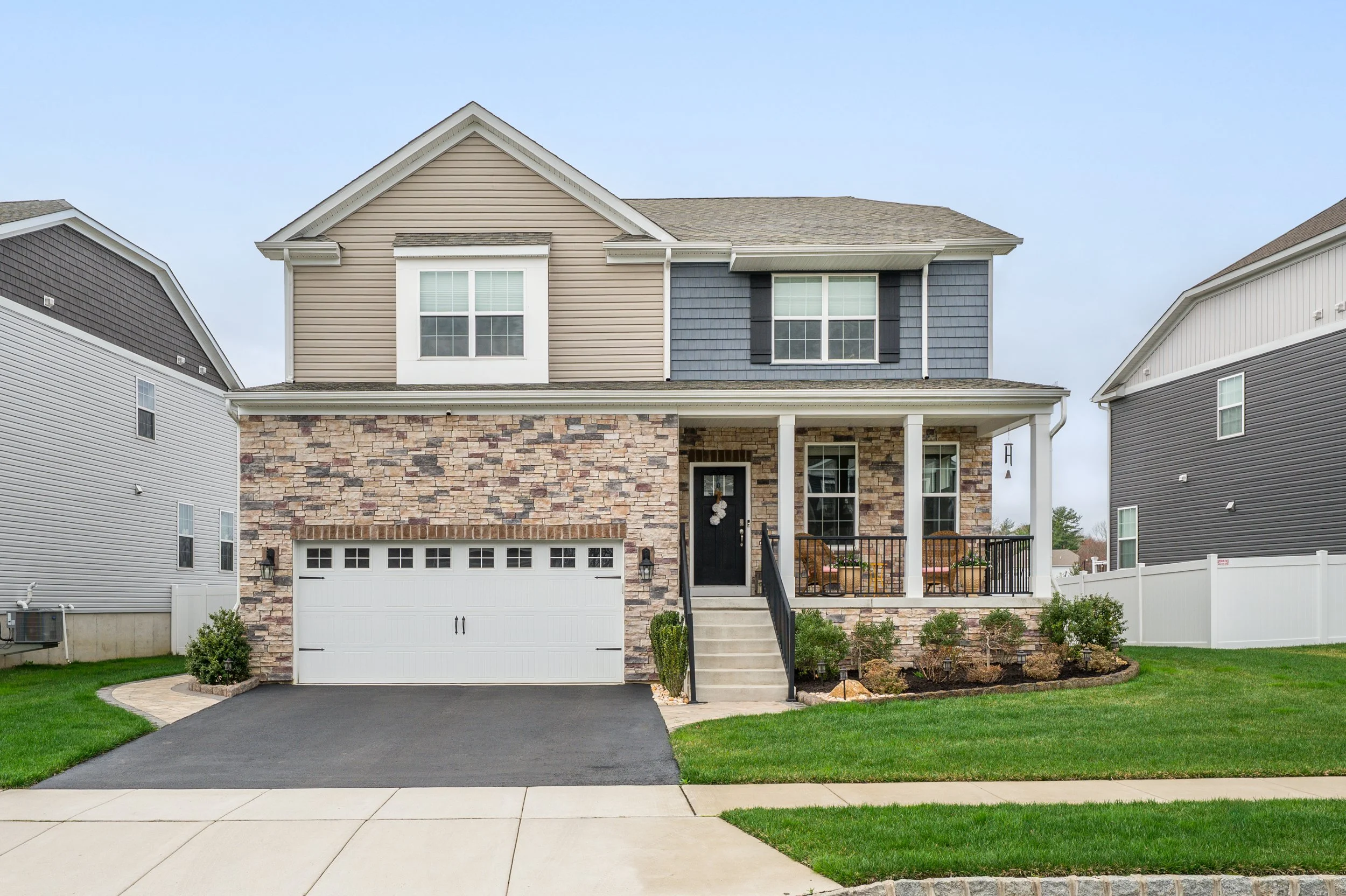 Front view of a modern two-story house with a stone and beige siding exterior, a white garage door, a small front porch with seating, and a well-maintained lawn.